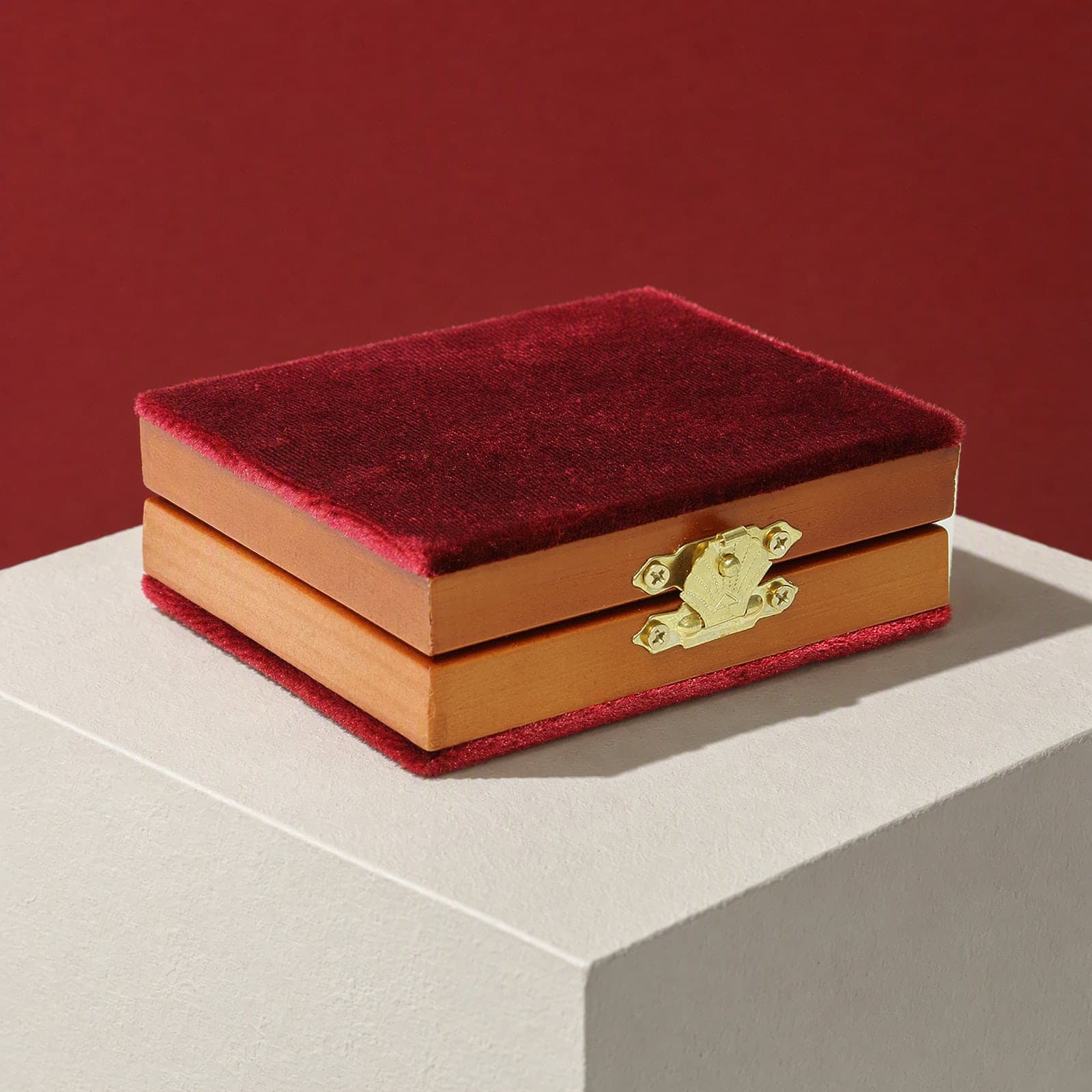 The folding diptych closed in its crimson velvet covers with honey pine edges and a brass clasp, on a white plinth against an oxblood backdrop.