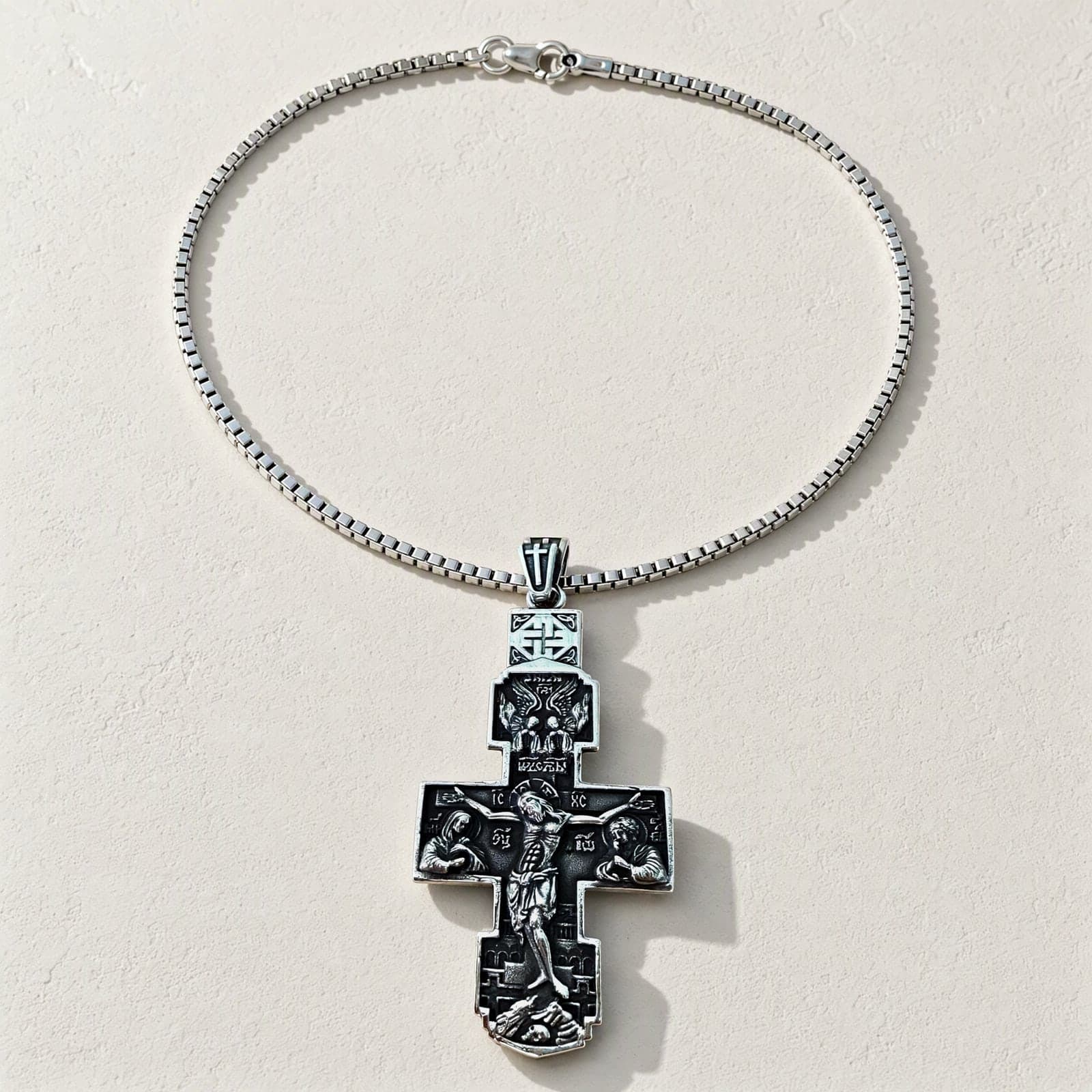Orthodox Crucifix - Silver Color - Stainless Steel Cross 20.2g laid flat with its plain silver-tone box chain on a white plaster surface.