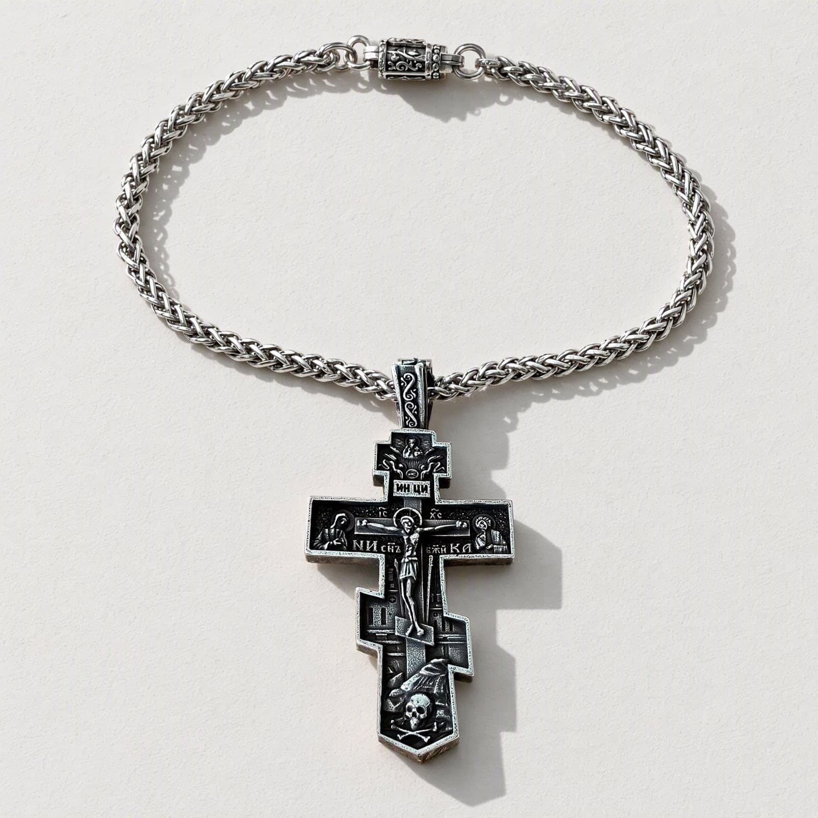Orthodox Crucifix (Mary and St John, Dark) - Silver Color - 60cm Chain - Stainless Steel Cross laid flat with its silver-tone wheat chain on a white plaster surface.