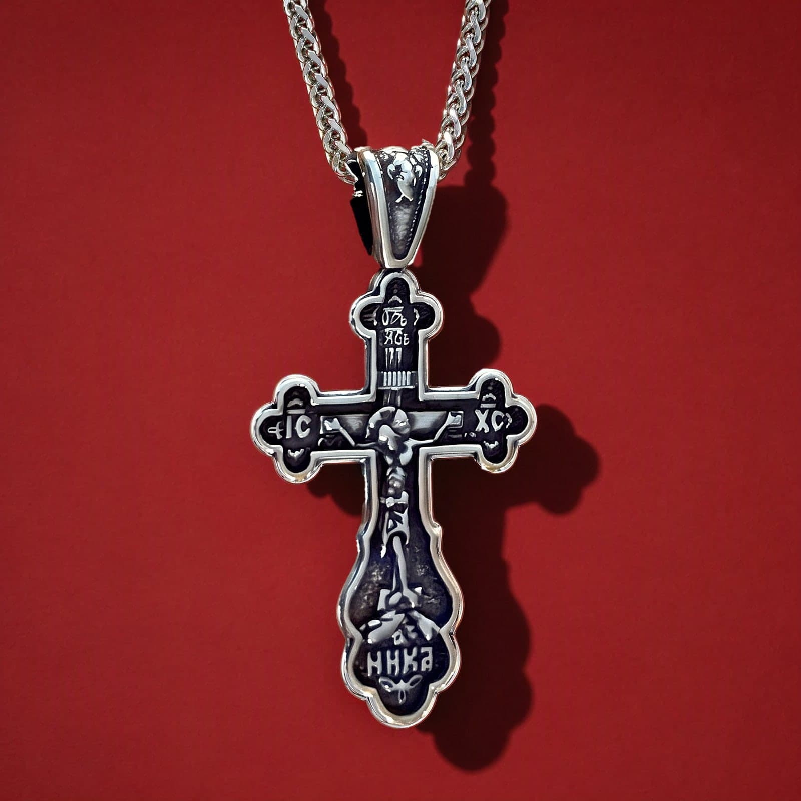 Orthodox Crucifix (Budded, Dark) - Silver Color - Stainless Steel Cross: a silver-tone stainless steel Orthodox crucifix with trefoil ends and deep relief on a silver-tone wheat chain, against an oxblood backdrop.