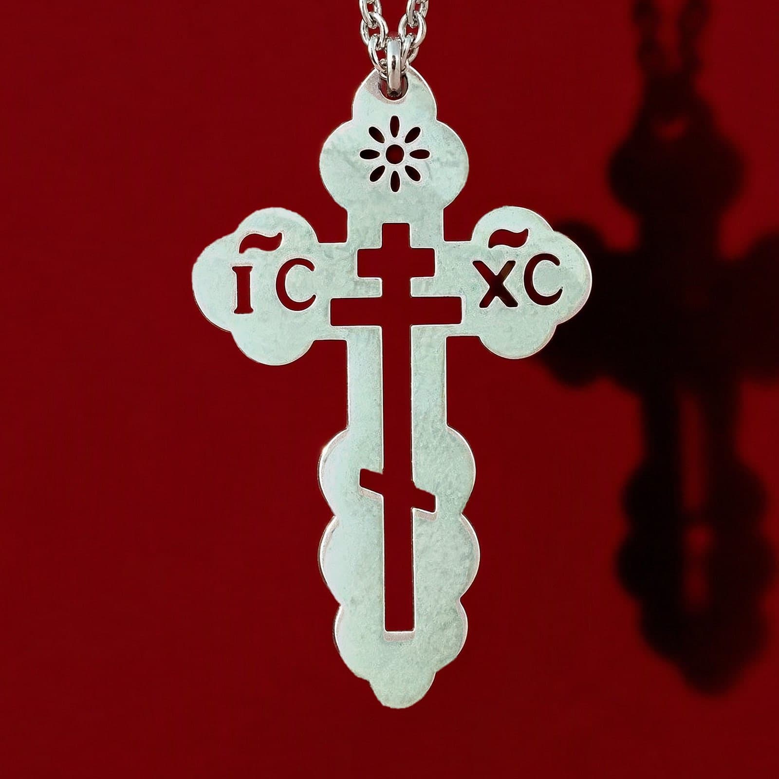 Orthodox IC XC - Silver Color - Stainless Steel Cross: a small silver-tone pierced Orthodox cross with trefoil ends, the IC XC letters and a cut-out three-bar cross on a fine silver-tone cable chain, against an oxblood backdrop.