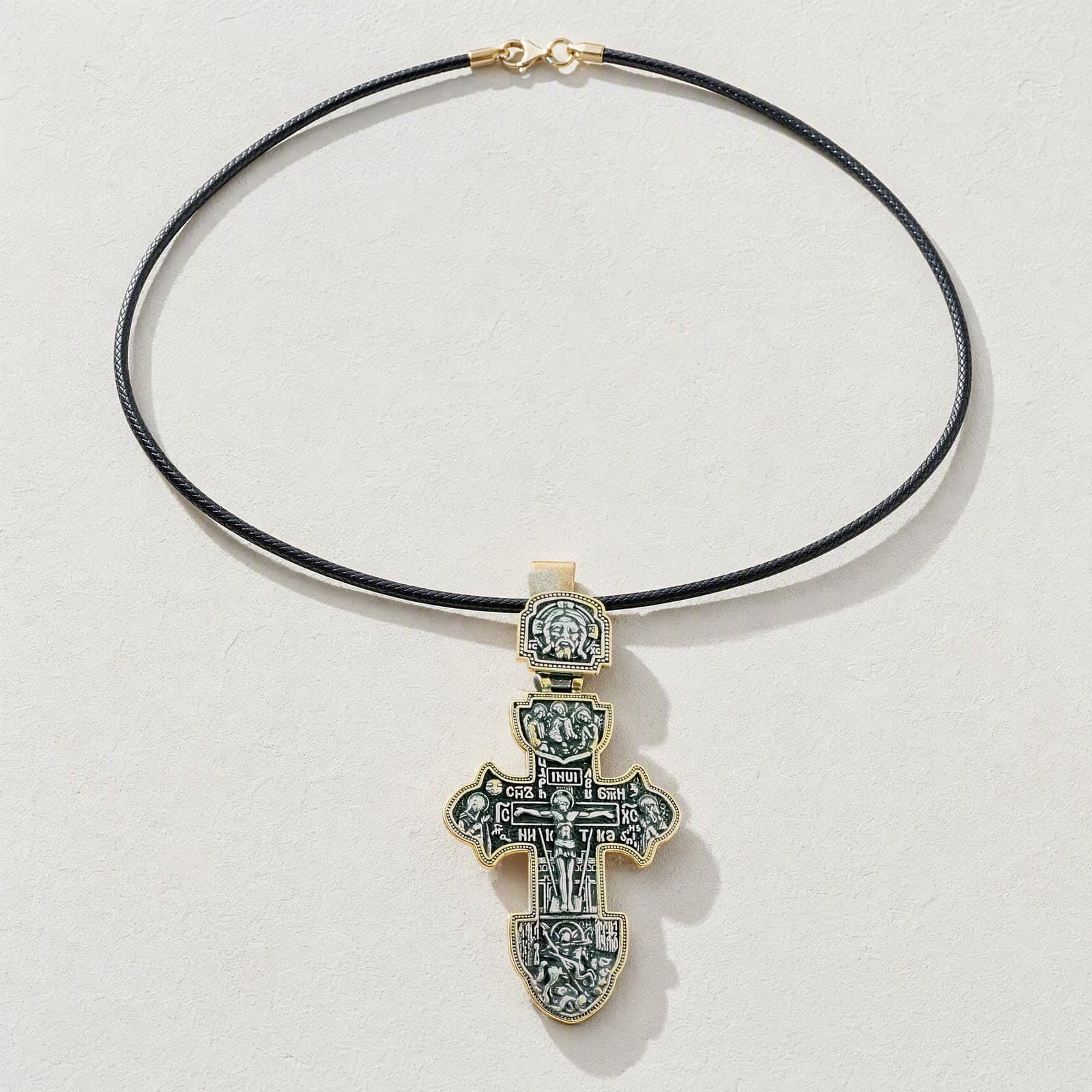 Orthodox Crucifix (Ornate) - Black&Gold Color - Stainless Steel Cross laid flat with its dark leather cord on a white plaster surface.