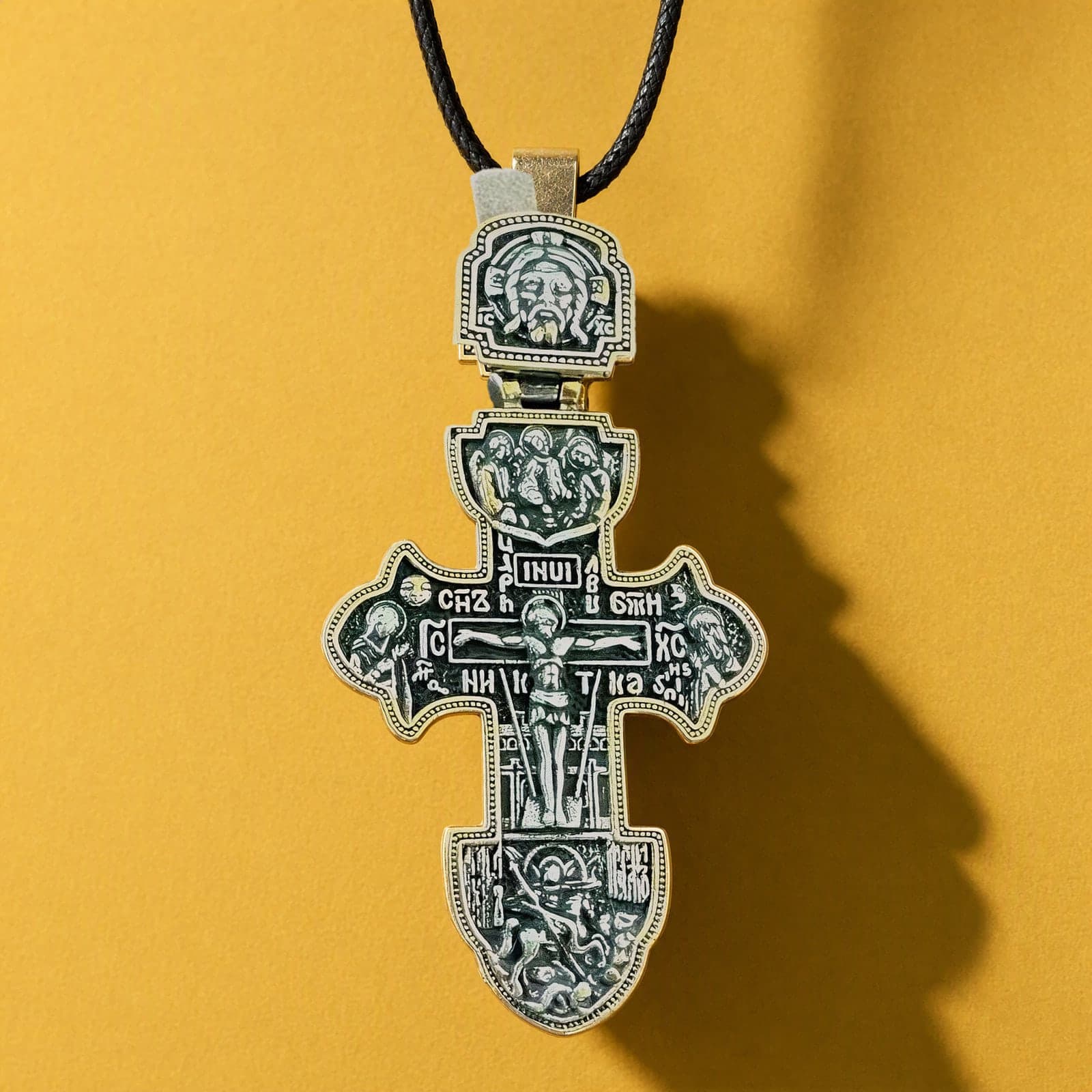 Orthodox Crucifix (Ornate) - Black&Gold Color - Stainless Steel Cross: a black-and-gold Orthodox crucifix with ornate relief and a gold-tone bail on a dark leather cord, against an ochre backdrop.
