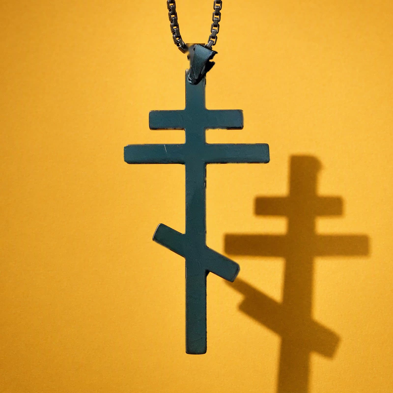 Orthodox Crucifix - Black Color - 60cm Chain - Stainless Steel Cross: a plain matte black stainless steel three-bar Orthodox cross with the slanted foot bar on a black box chain, against an ochre backdrop.