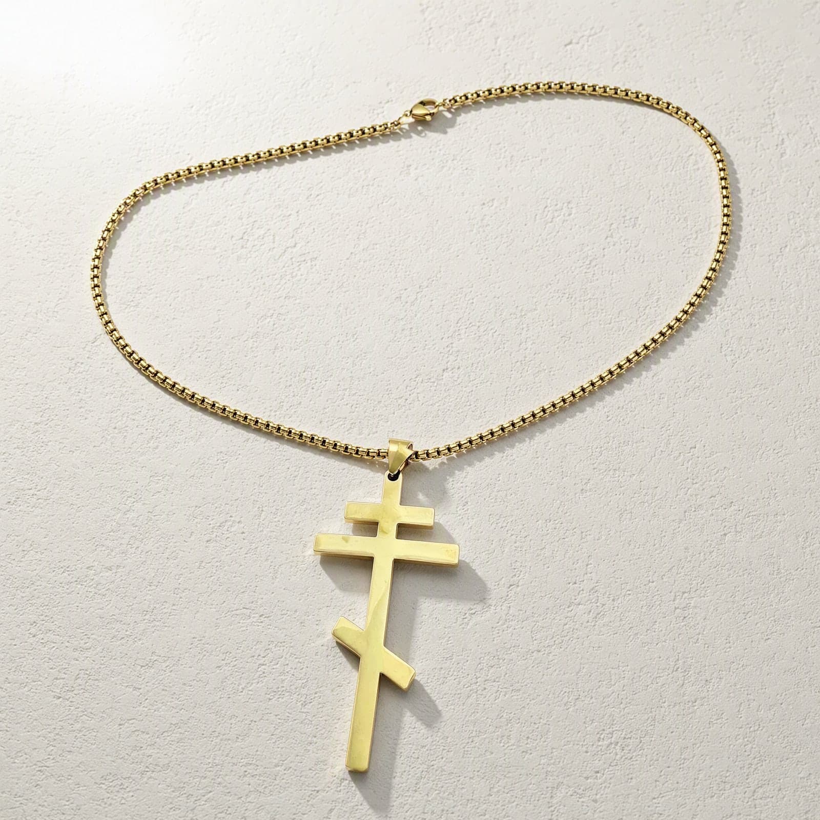 Orthodox Crucifix - Gold Color - 60cm Chain - Stainless Steel Cross laid flat with its gold-tone box chain on a white plaster surface.