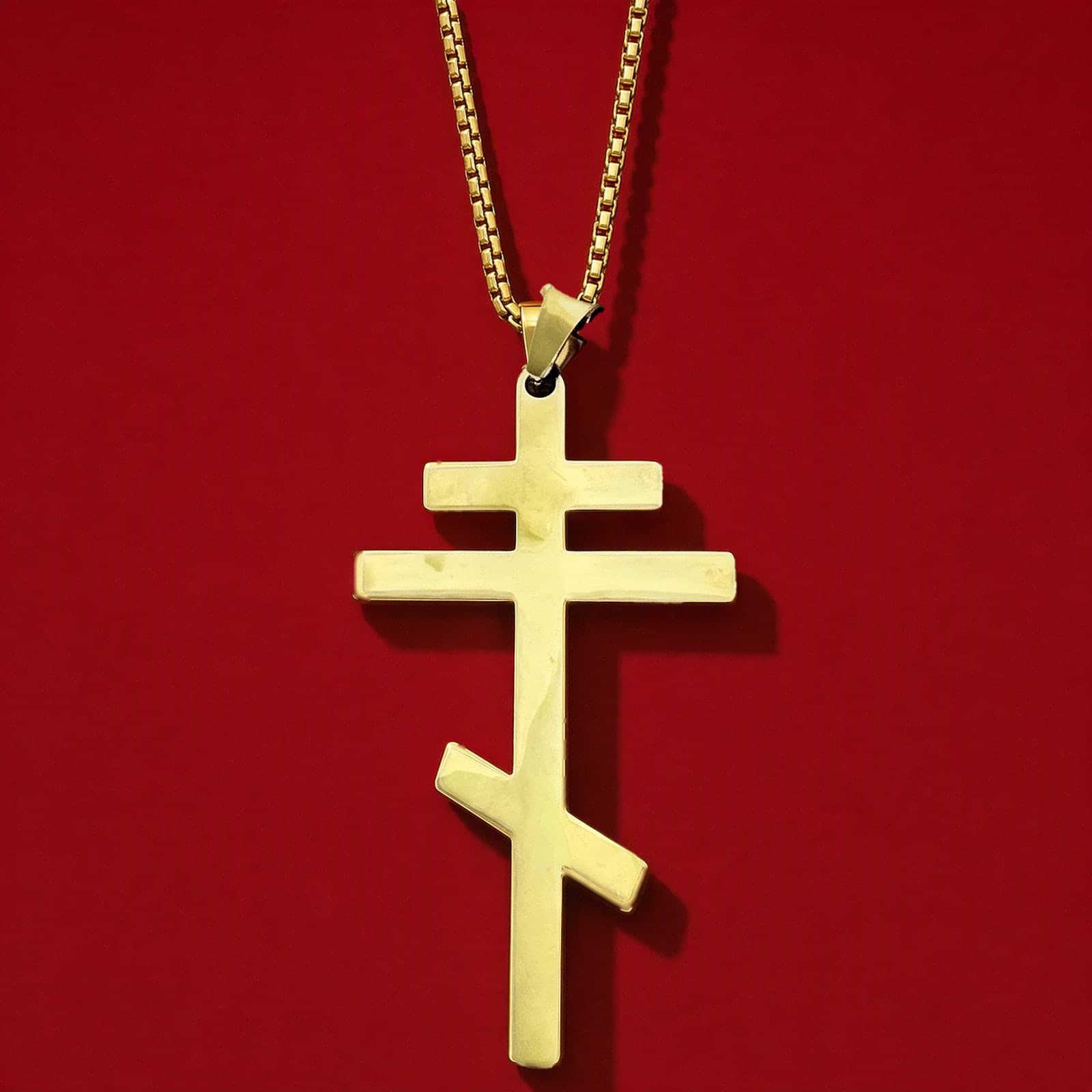 Orthodox Crucifix - Gold Color - 60cm Chain - Stainless Steel Cross: a plain polished gold-tone stainless steel three-bar Orthodox cross with the slanted foot bar on a gold-tone box chain, against an oxblood backdrop.