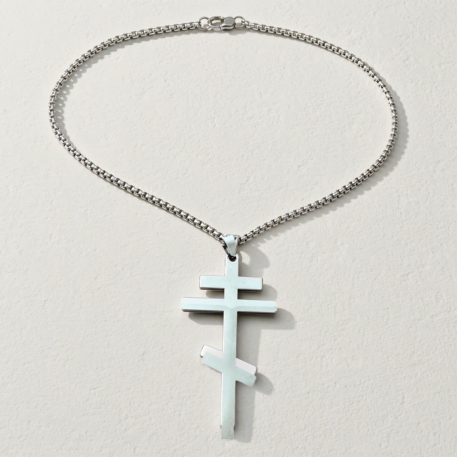 Orthodox Crucifix (Plain Three-Bar) - Silver Color - 60cm Chain - Stainless Steel Cross laid flat with its silver-tone box chain on a white plaster surface.
