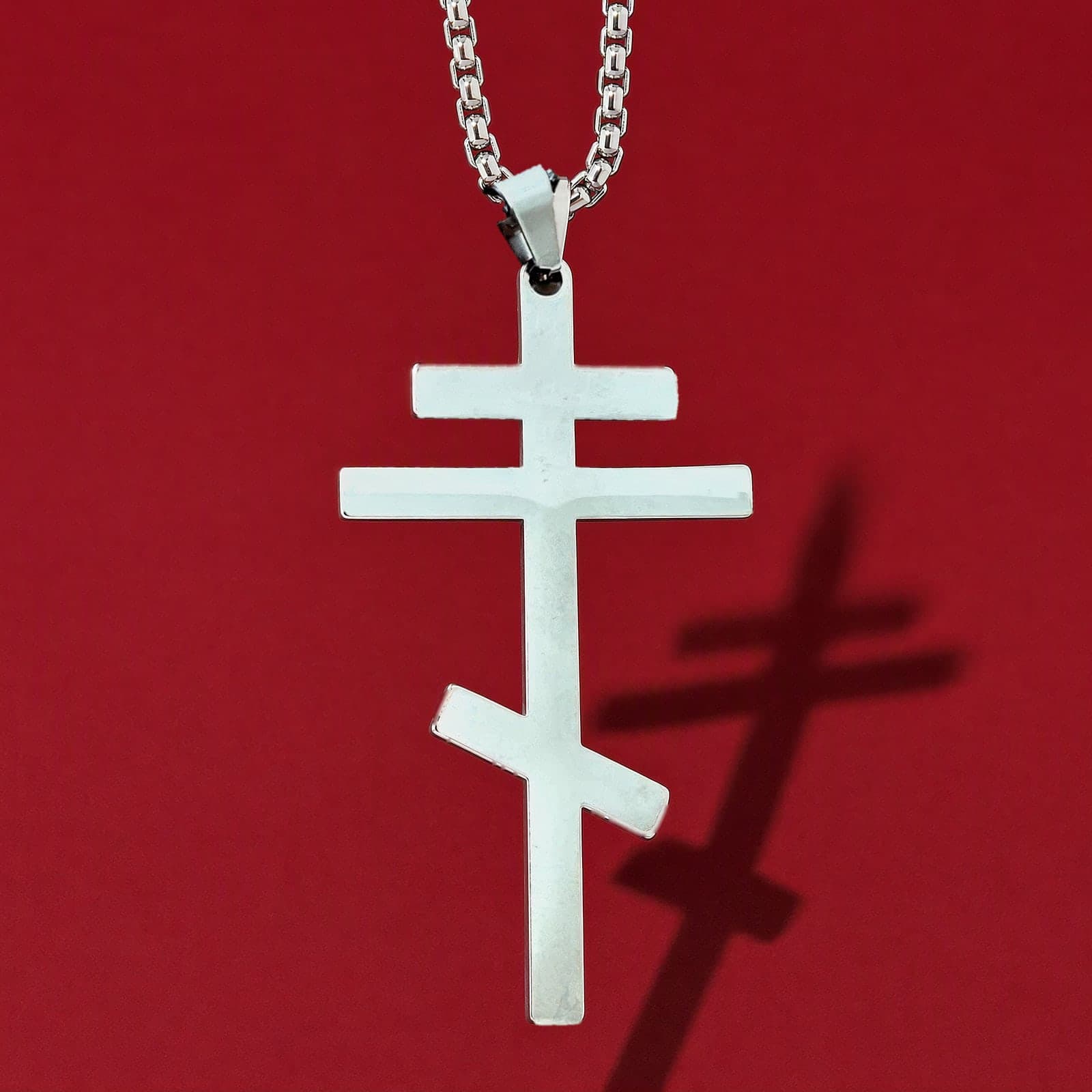 Orthodox Crucifix (Plain Three-Bar) - Silver Color - 60cm Chain - Stainless Steel Cross: a plain polished silver-tone stainless steel three-bar Orthodox cross with the slanted foot bar on a silver-tone box chain, against an oxblood backdrop.