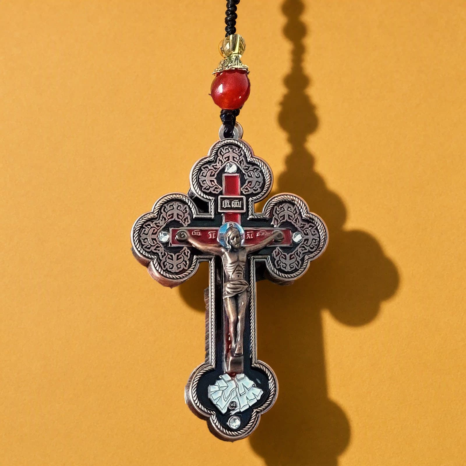 Orthodox Crucifix - Copper Red Color - Stainless Steel Cross: a copper-red enamelled Orthodox crucifix with trefoil ends and a red agate bead above it on a black knotted cord, against an ochre backdrop.