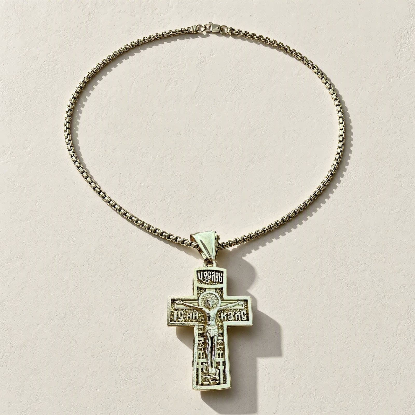 Orthodox Crucifix (King of Glory) - Gold Color - Stainless Steel Cross laid flat with its gold-tone box chain on a white plaster surface.