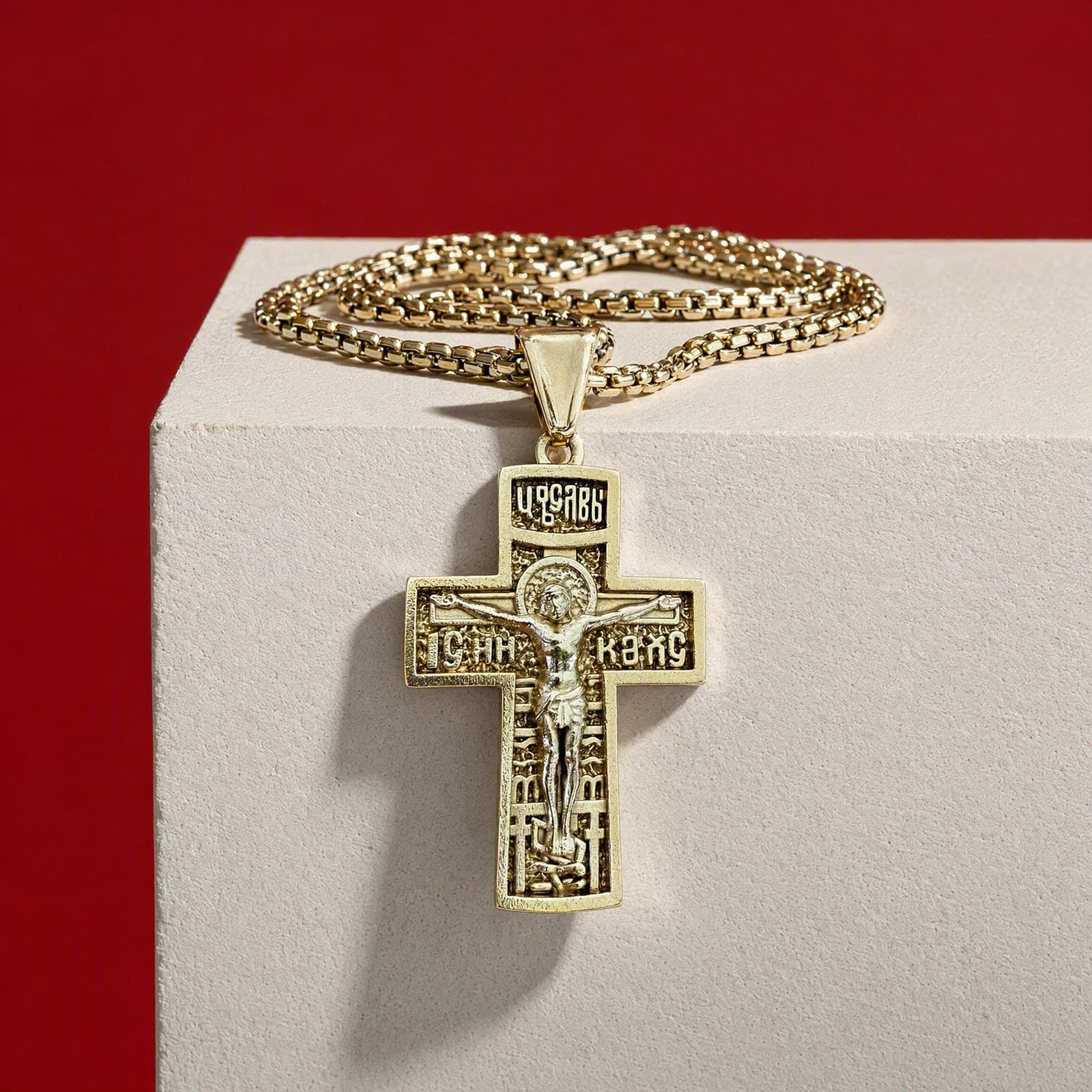 Orthodox Crucifix (King of Glory), gold-tone stainless steel cross, hanging over the edge of a bone plaster plinth against an oxblood backdrop, on a gold-tone box chain.