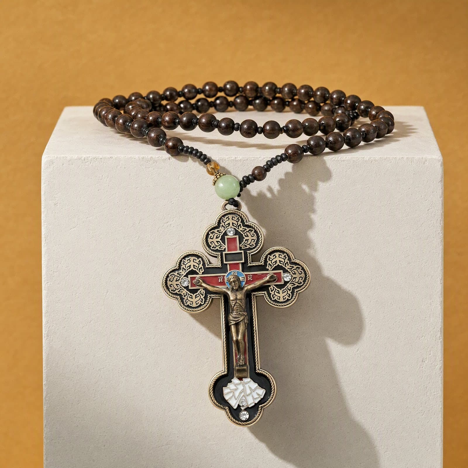 Orthodox Crucifix, bronze-tone cross, hanging over the edge of a bone plaster plinth against an ochre backdrop, on a strand of dark brown wooden beads that ends in a short knotted black cord with a pale jade bead just above the cross.