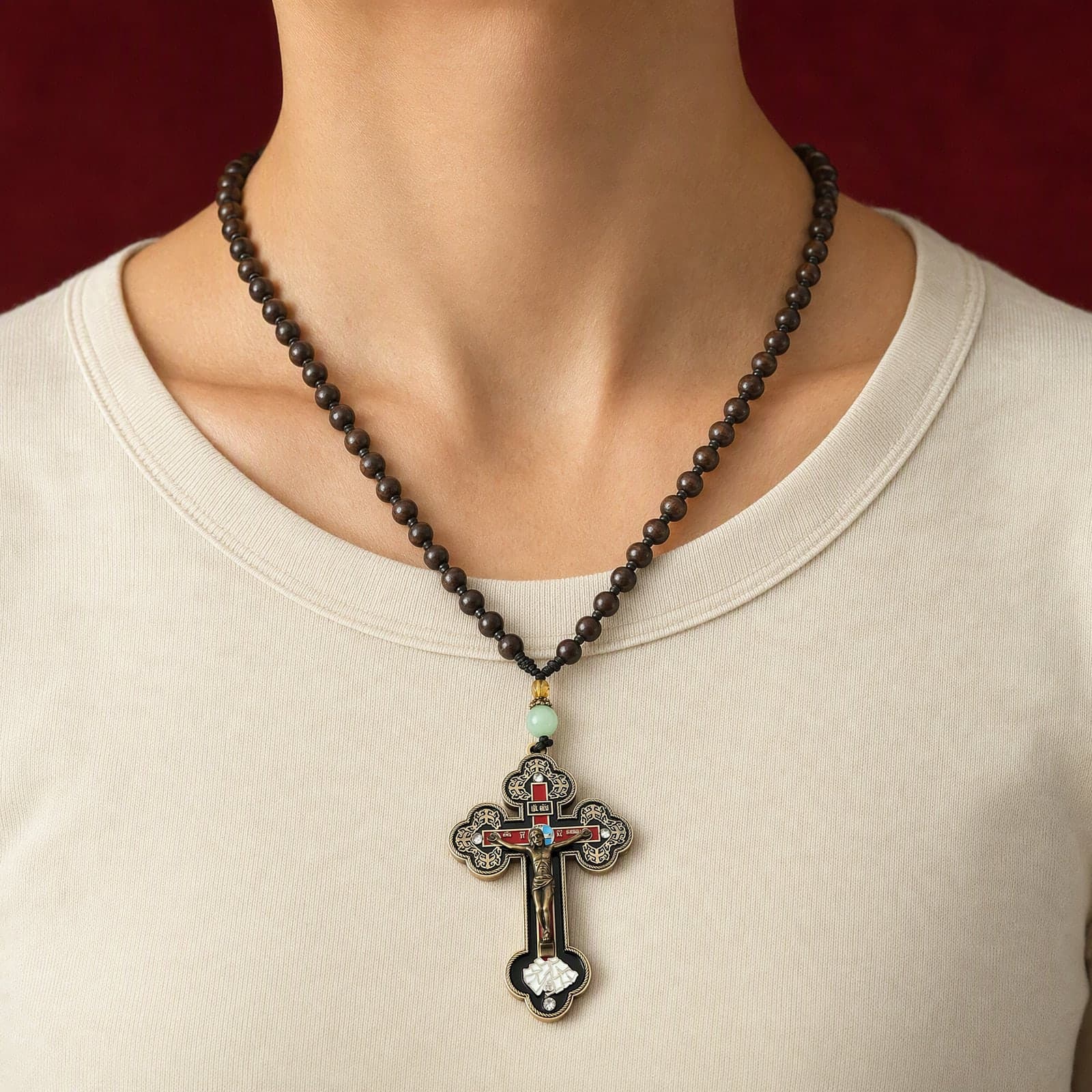 Orthodox Crucifix, bronze-tone cross worn at its true size (5.5 cm) on a strand of dark brown wooden beads that ends in a short knotted black cord with a pale jade bead just above the cross, close crop of the chest.