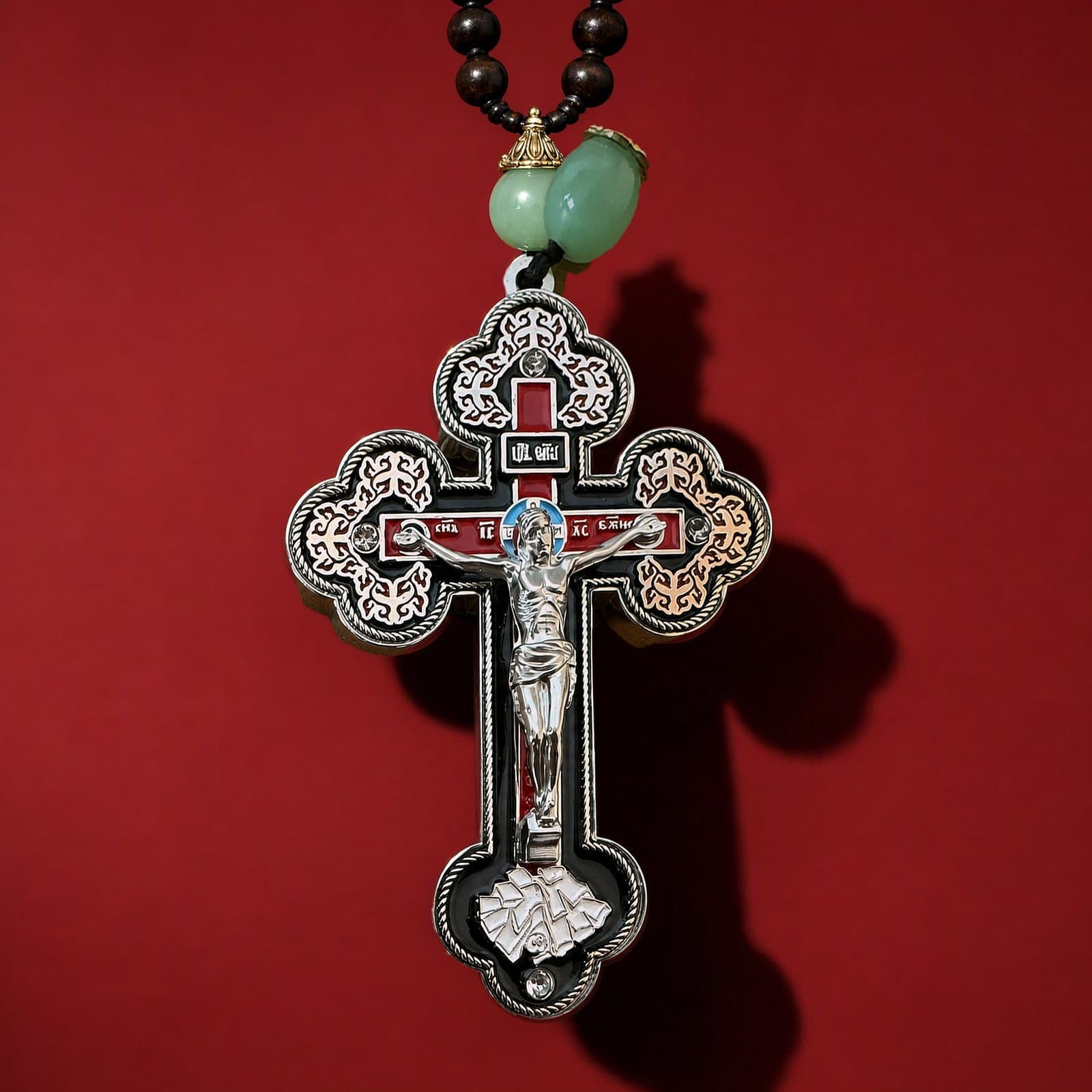 Orthodox Crucifix - Silver Color - Cross: a silver-tone enamelled Orthodox crucifix with trefoil ends, hung from a pale jade bead on a dark bead chain, against an oxblood backdrop.