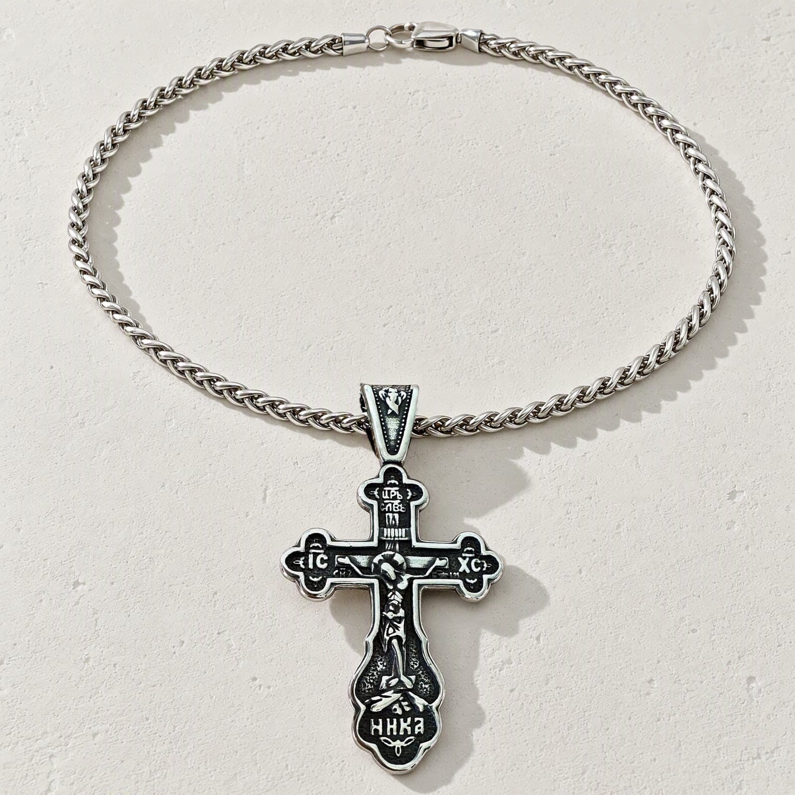 Orthodox Crucifix (Budded, Bright) - Silver Color - Stainless Steel Cross laid flat with its silver-tone wheat chain on a white plaster surface.