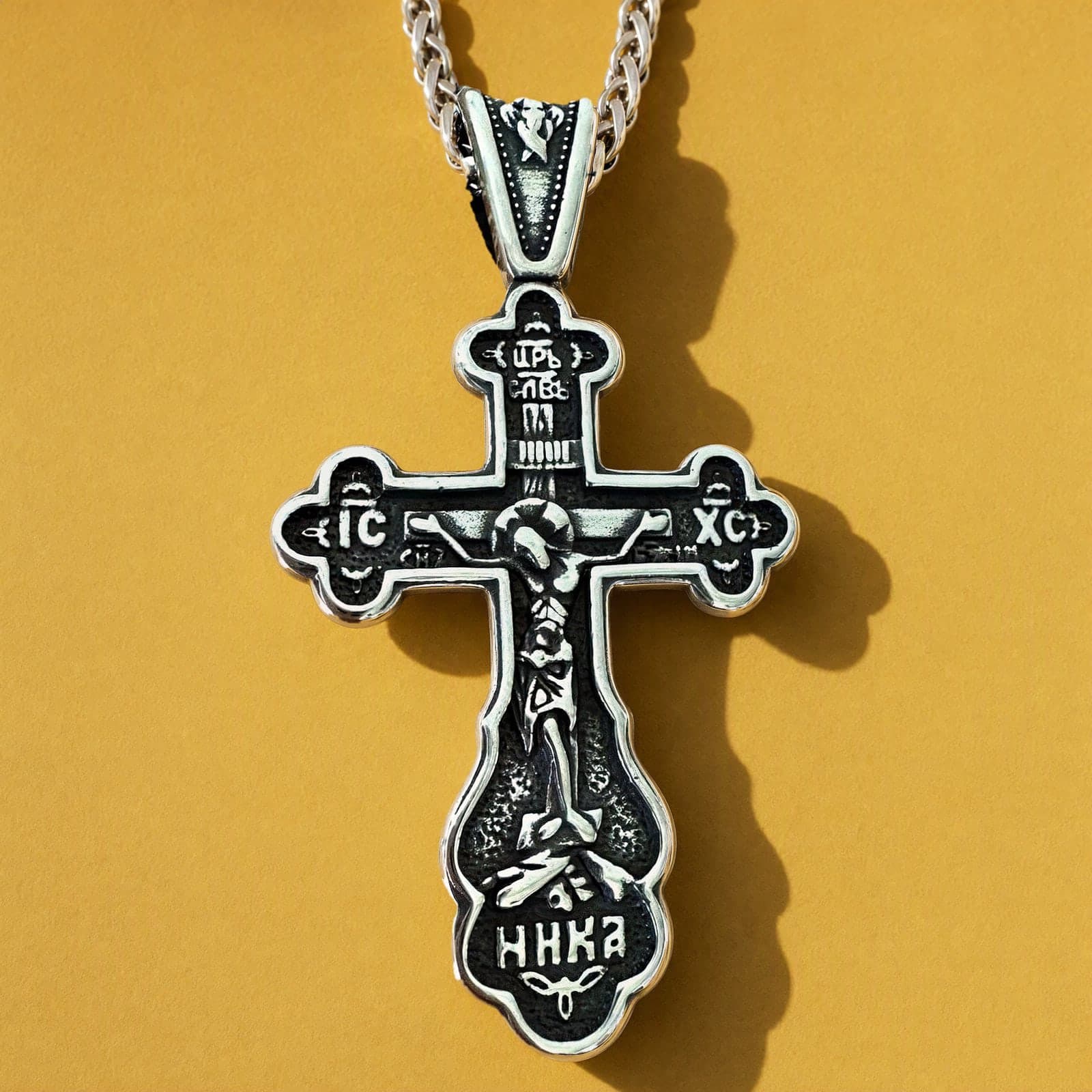 Orthodox Crucifix (Budded, Bright) - Silver Color - Stainless Steel Cross: a blackened silver-tone Orthodox crucifix with trefoil ends and deep relief on a silver-tone wheat chain, against an ochre backdrop.