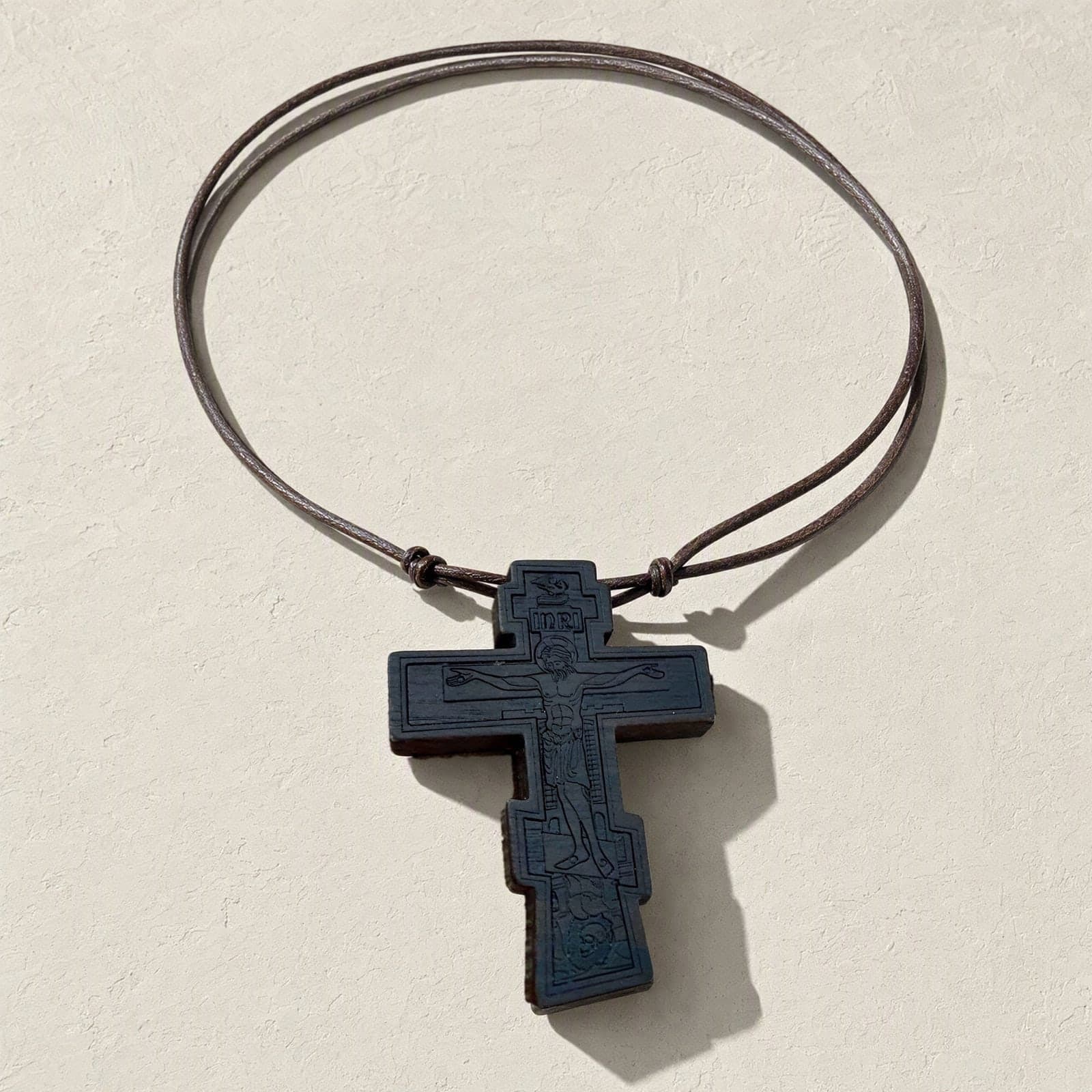 Orthodox Crucifix - Black Color - Wooden Cross laid flat with its thin brown leather cord on a white plaster surface.