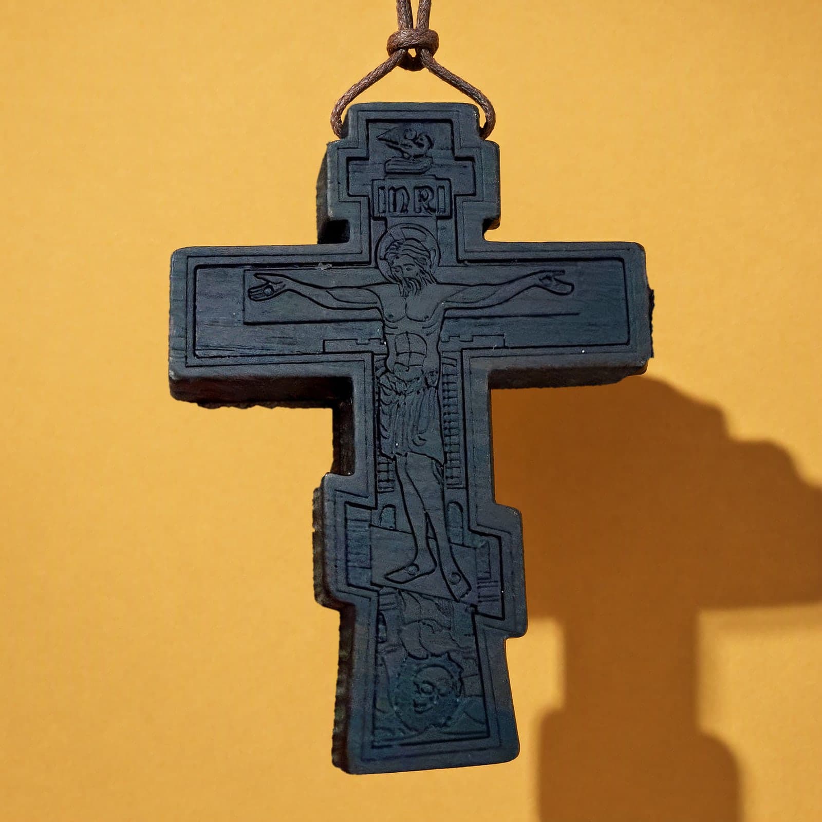 Orthodox Crucifix - Black Color - Wooden Cross: a black-stained wooden Orthodox crucifix with carved relief on a thin brown leather cord, against an ochre backdrop.