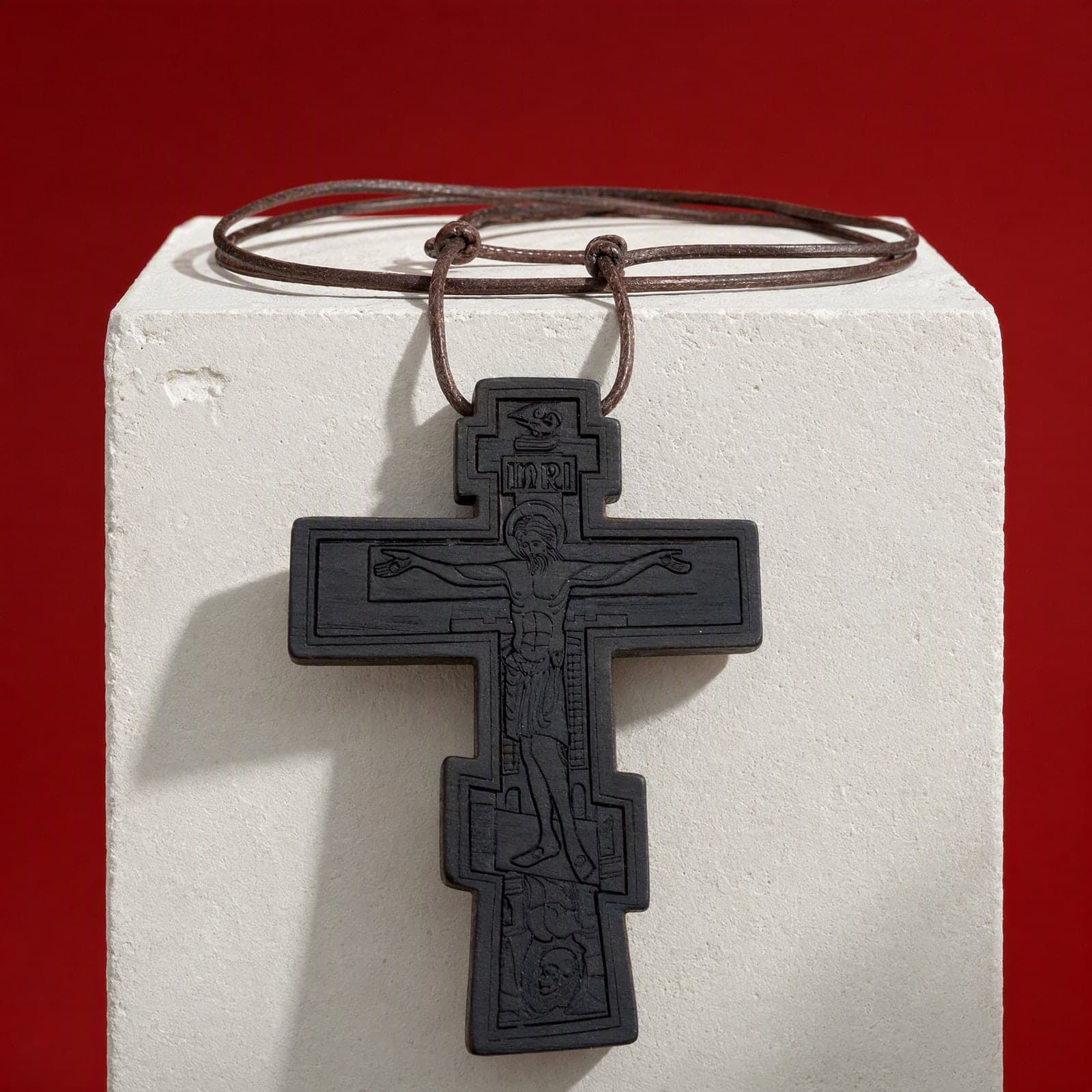 Православный крест с распятием, чёрный, деревянный — свисает с края гипсовой подставки цвета слоновой кости на тёмно-бордовом фоне, на тонком коричневом кожаном шнурке.
