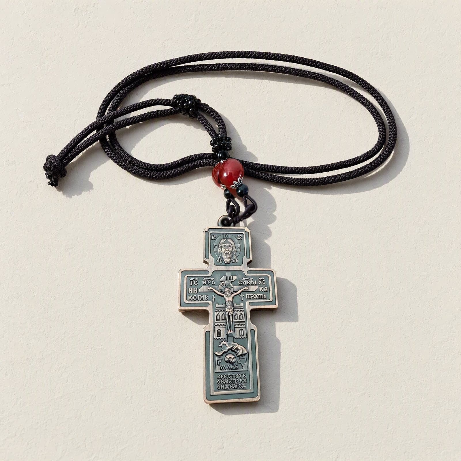 Orthodox Crucifix (Holy Face, Copper) - Bronze Color - Stainless Steel Cross laid flat with its dark knotted cord on a white plaster surface.