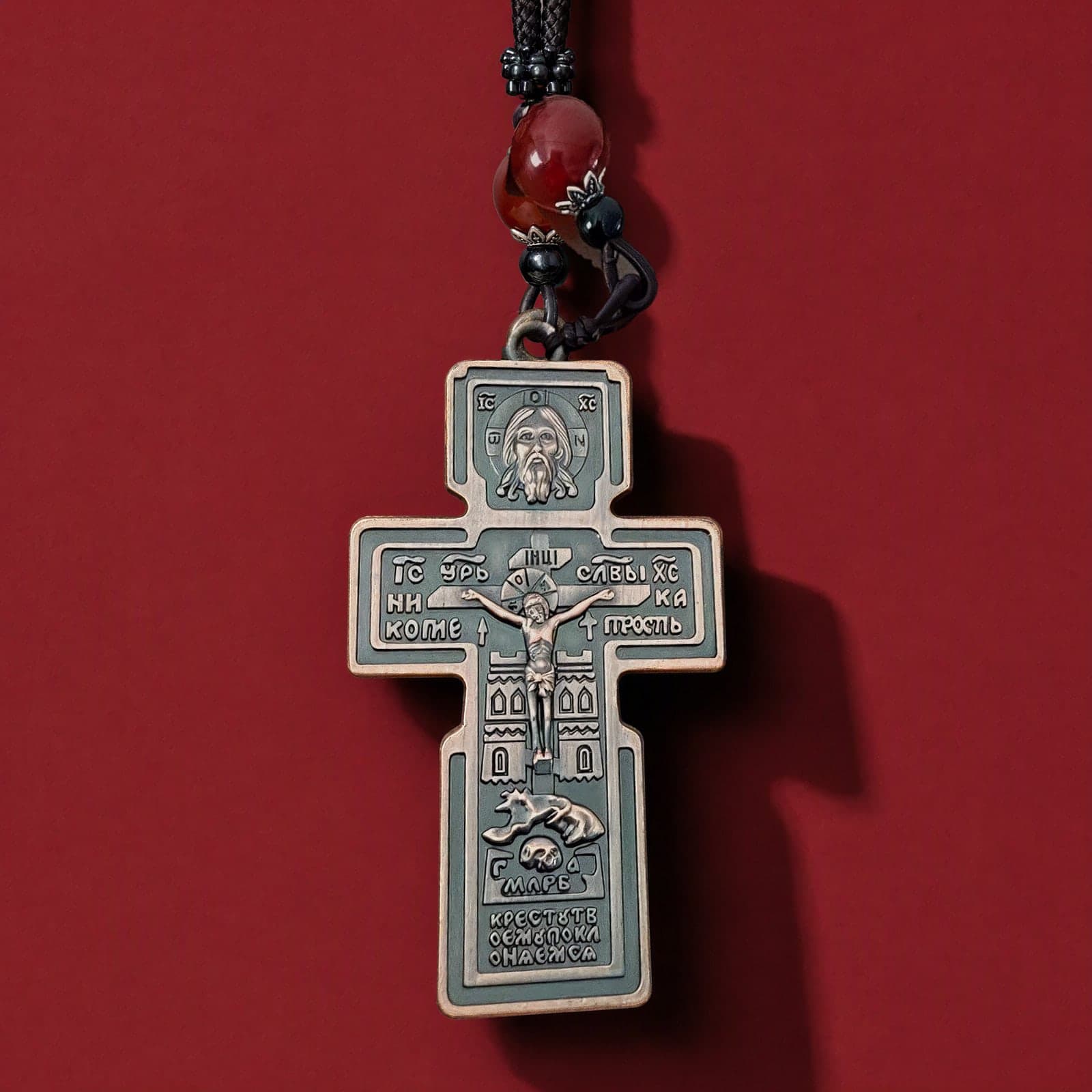 Orthodox Crucifix (Holy Face, Copper) - Bronze Color - Stainless Steel Cross: a bronze-tone Orthodox crucifix with deep relief, hung with a red agate bead on a dark knotted cord, against an oxblood backdrop.