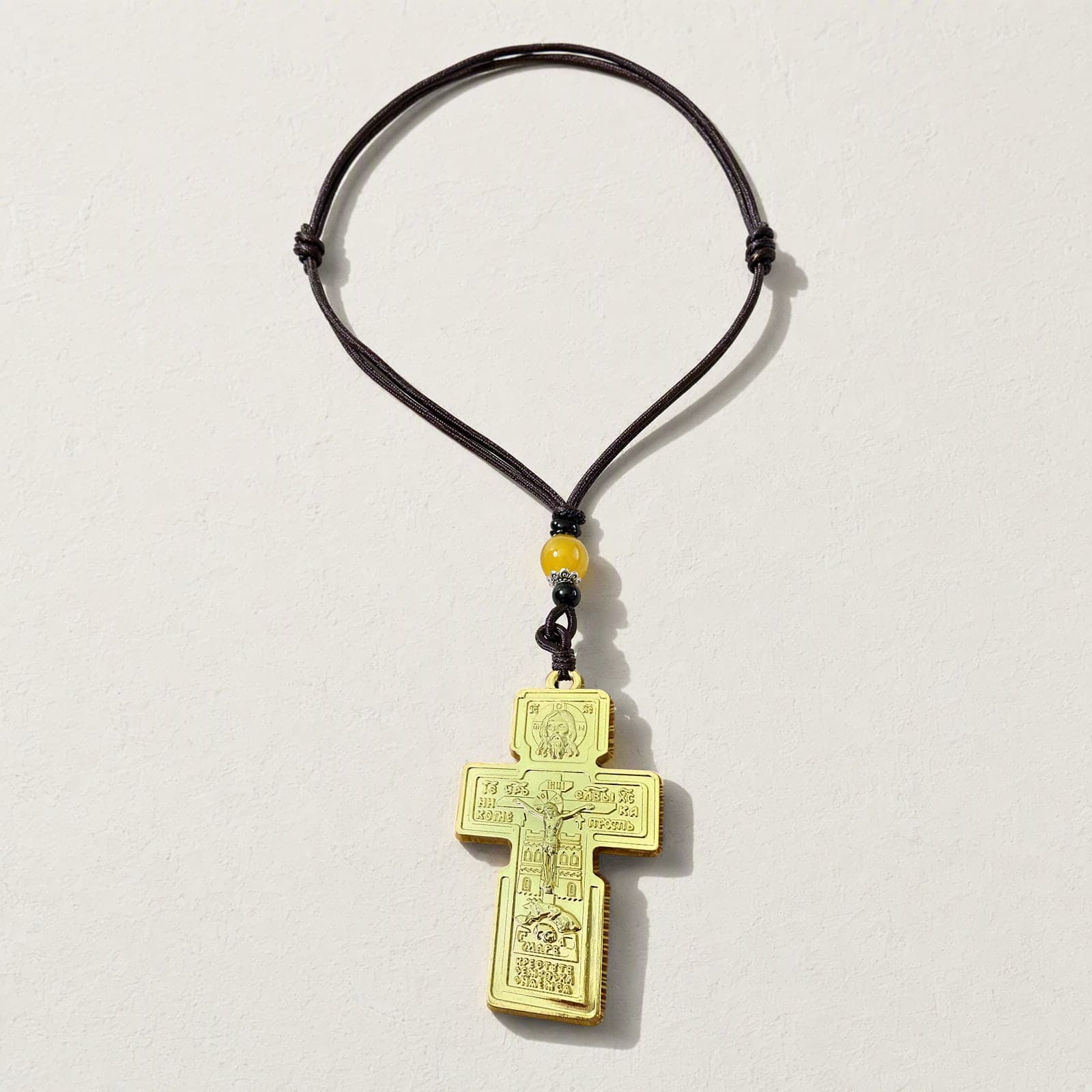 Orthodox Crucifix (Holy Face) - Gold Color - Stainless Steel Cross laid flat with its dark brown cord on a white plaster surface.