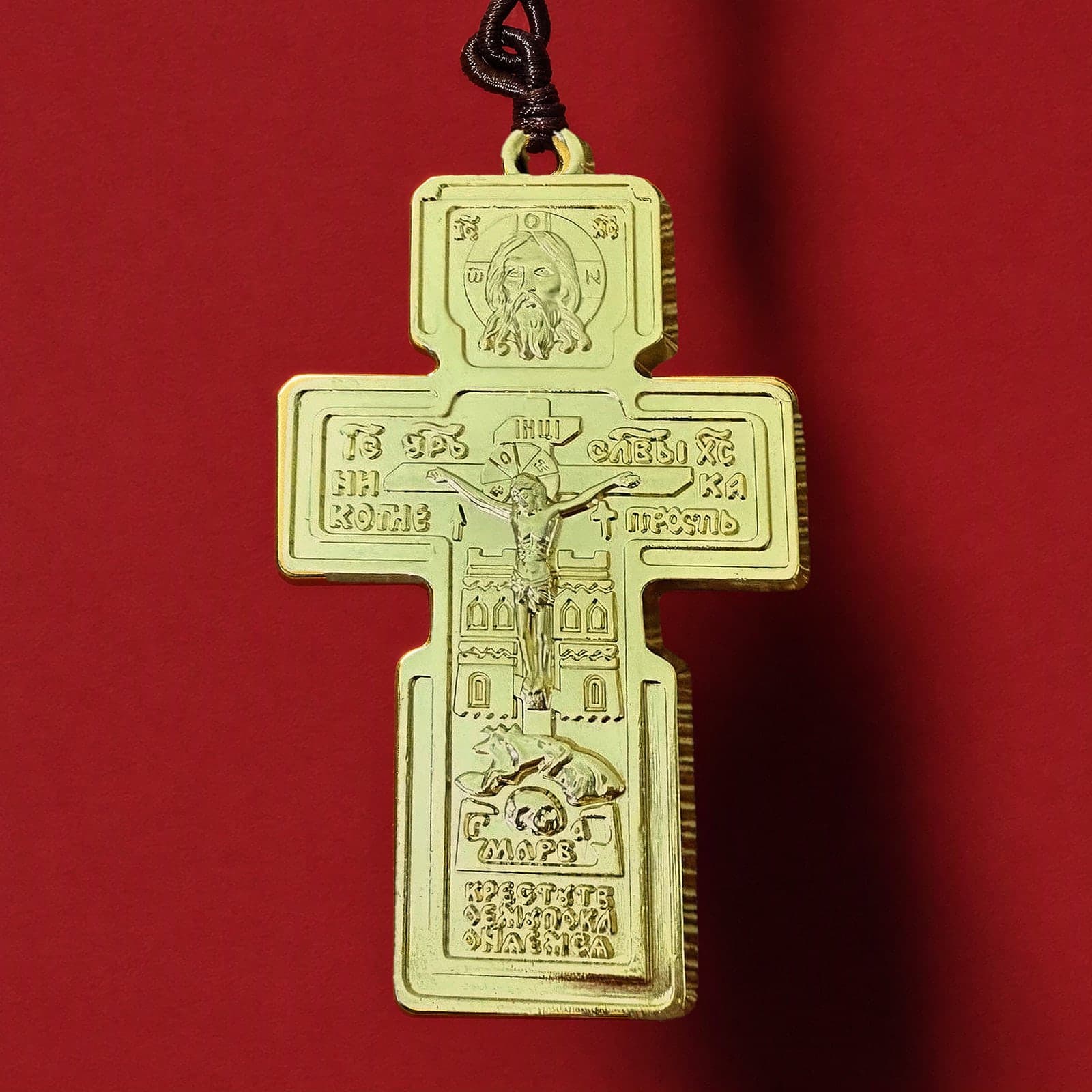 Orthodox Crucifix (Holy Face) - Gold Color - Stainless Steel Cross: a gold-tone Orthodox crucifix with lettered relief, hung with a yellow bead on a dark brown cord, against an oxblood backdrop.