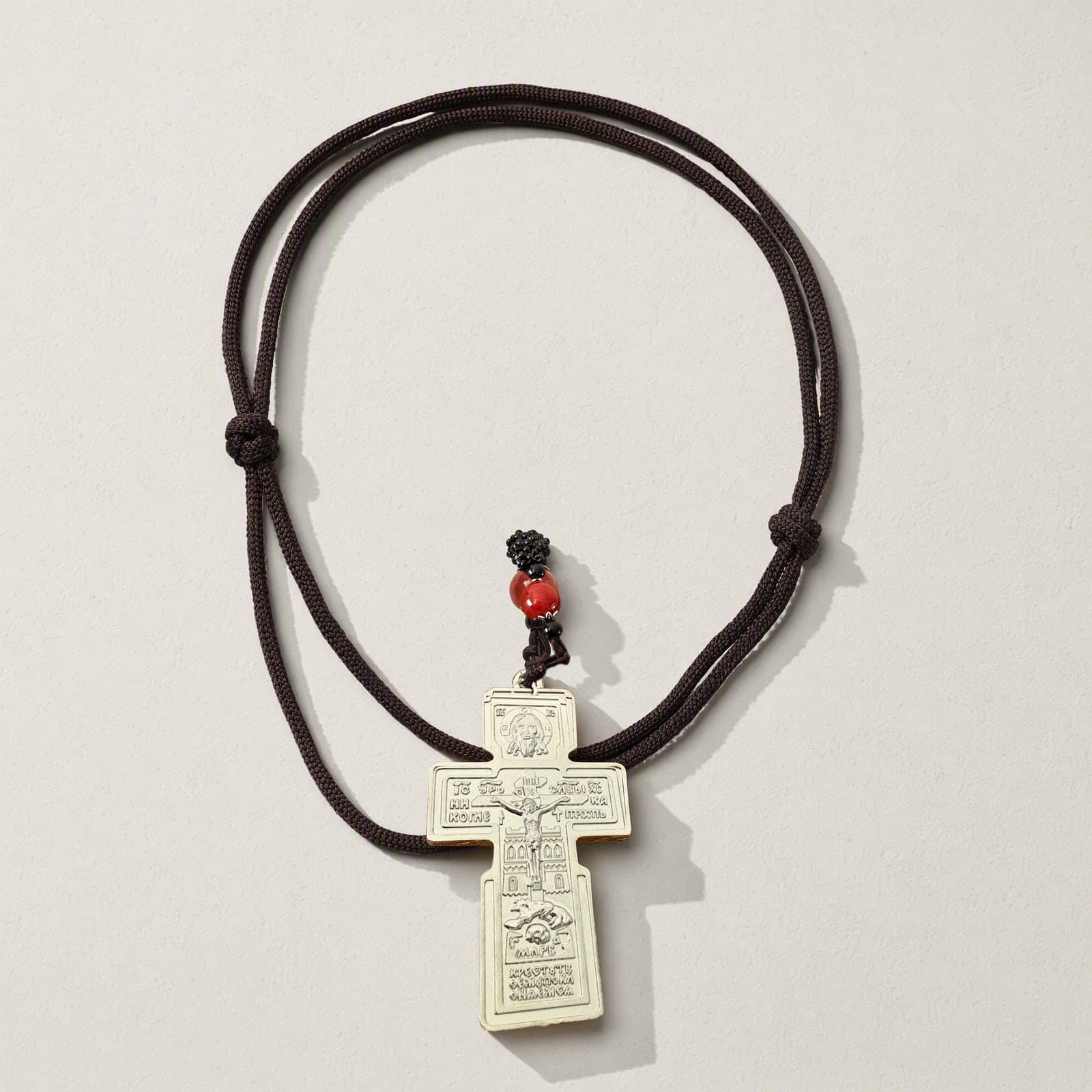 Orthodox Crucifix (Holy Face) - Silver Color - Stainless Steel Cross laid flat with its dark knotted cord on a white plaster surface.