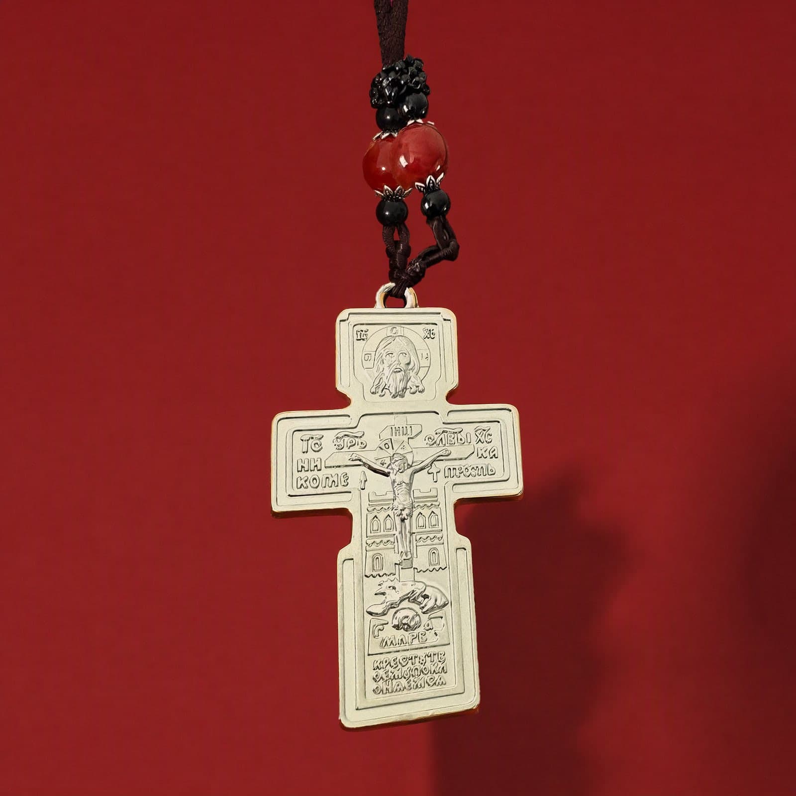 Orthodox Crucifix (Holy Face) - Silver Color - Stainless Steel Cross: a pale gold-tone Orthodox crucifix with lettered relief, hung with a red bead on a dark knotted cord, against an oxblood backdrop.