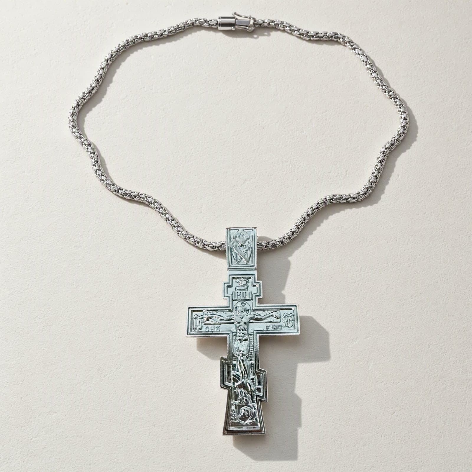Orthodox Crucifix (Son of God) - Silver Color - Stainless Steel Cross laid flat with its silver-tone box chain on a white plaster surface.