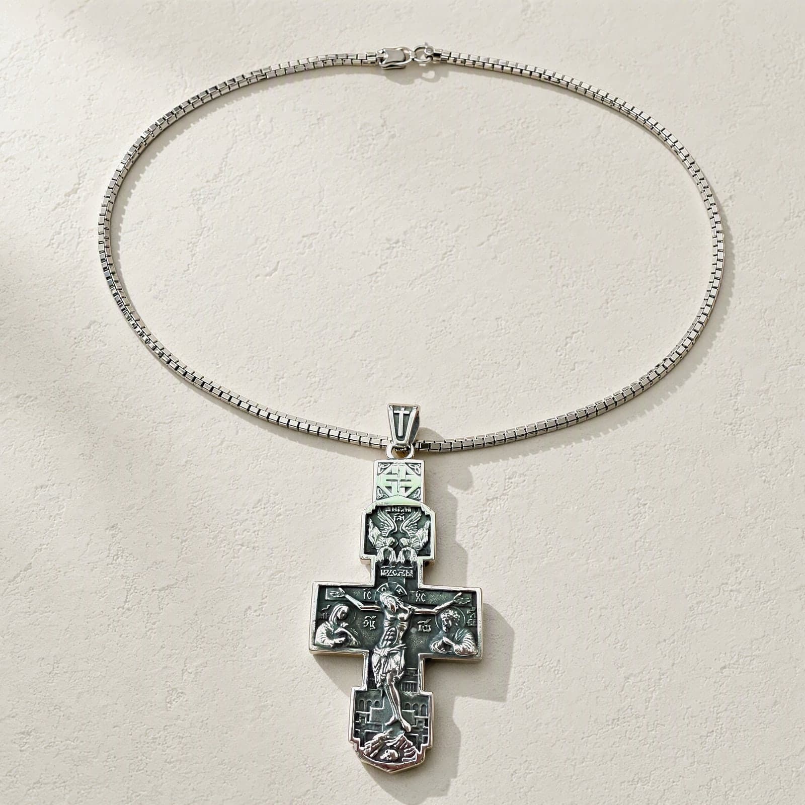 Orthodox Crucifix (Angels) - Silver Color - Stainless Steel Cross 70x39mm 20.2g laid flat with its plain silver-tone box chain on a white plaster surface.