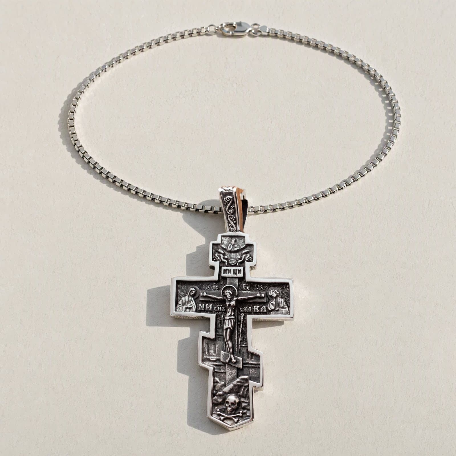 Orthodox Crucifix (Mary and St John, Bright) - Silver Color - Stainless Steel Cross 70x39mm 20.2g laid flat with its plain silver-tone box chain on a white plaster surface.