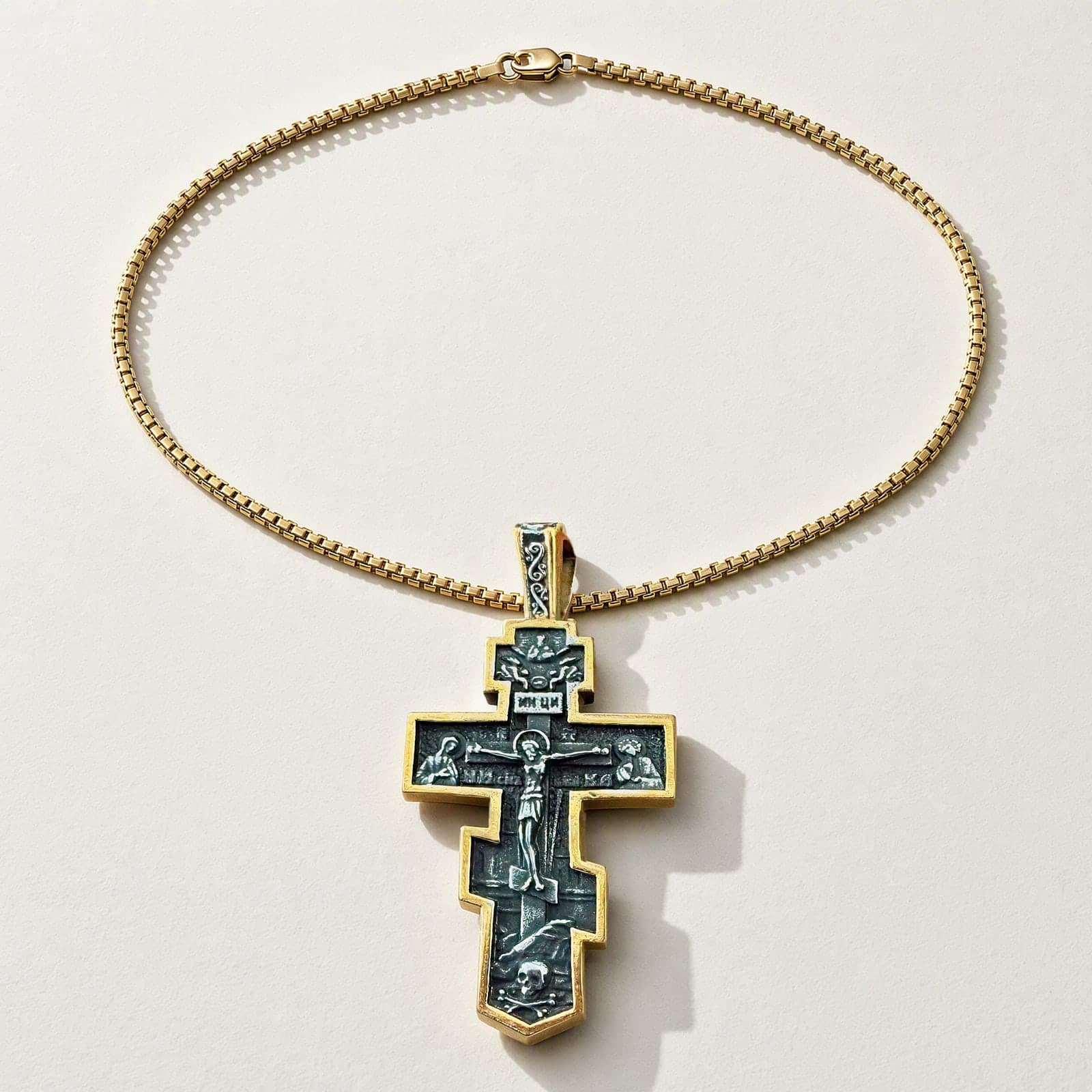 Orthodox Crucifix (Mary and St John) - Black&Gold Color - Stainless Steel Cross 70x39mm 20.2g laid flat with its plain gold-tone box chain on a white plaster surface.