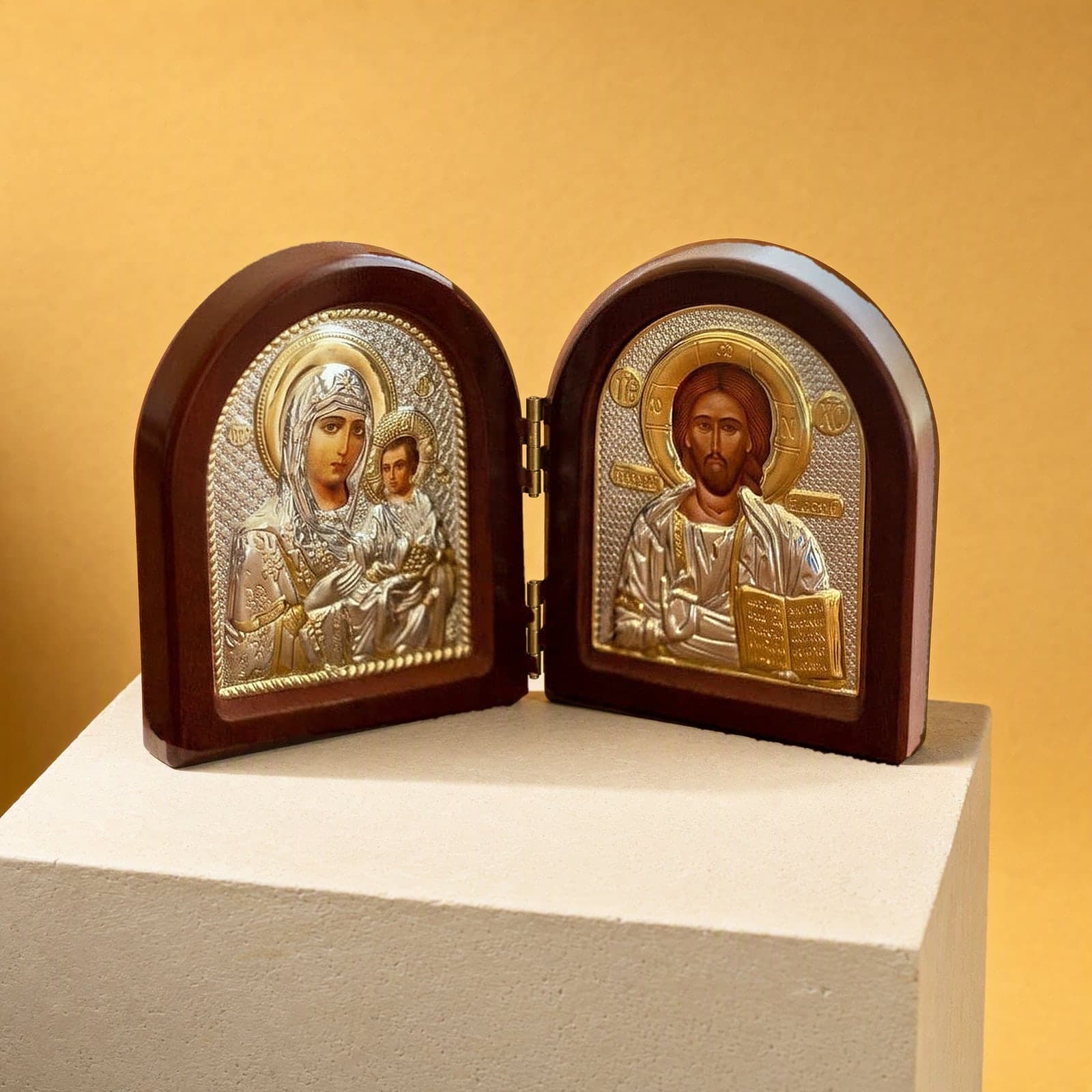 Orthodox Jesus Christ and Virgin Mary - Folding Wooden Icon: a small folding arched icon diptych with silver-and-gold foil icons of Christ and the Mother of God in dark wooden frames, on a white plinth against an ochre backdrop.