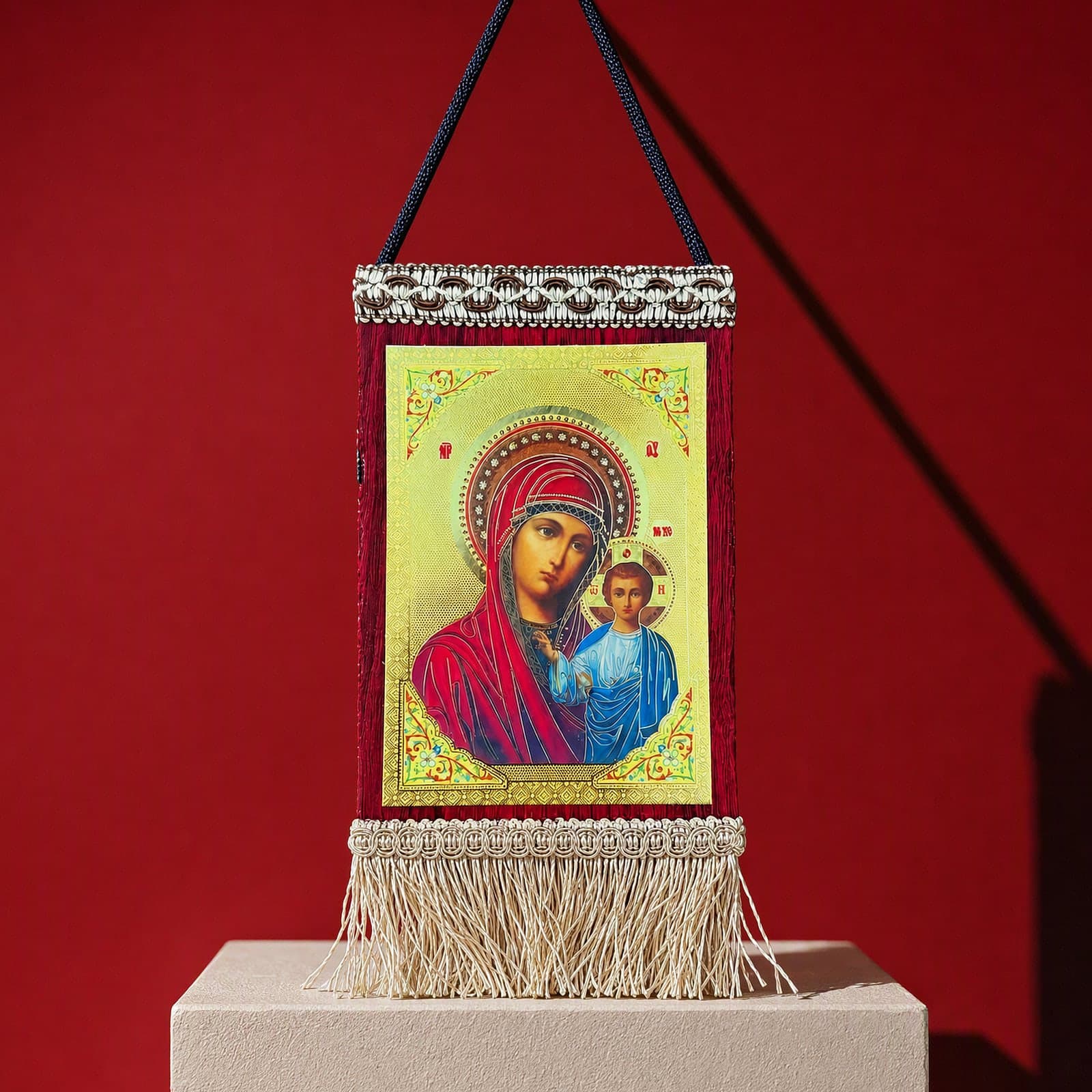 Orthodox Virgin Mary & Jesus - Hanging Icon 20x13cm: a small textile banner icon of the Mother of God with the Christ Child with a cream fringe and a hanging cord, on a white plinth against an oxblood backdrop.