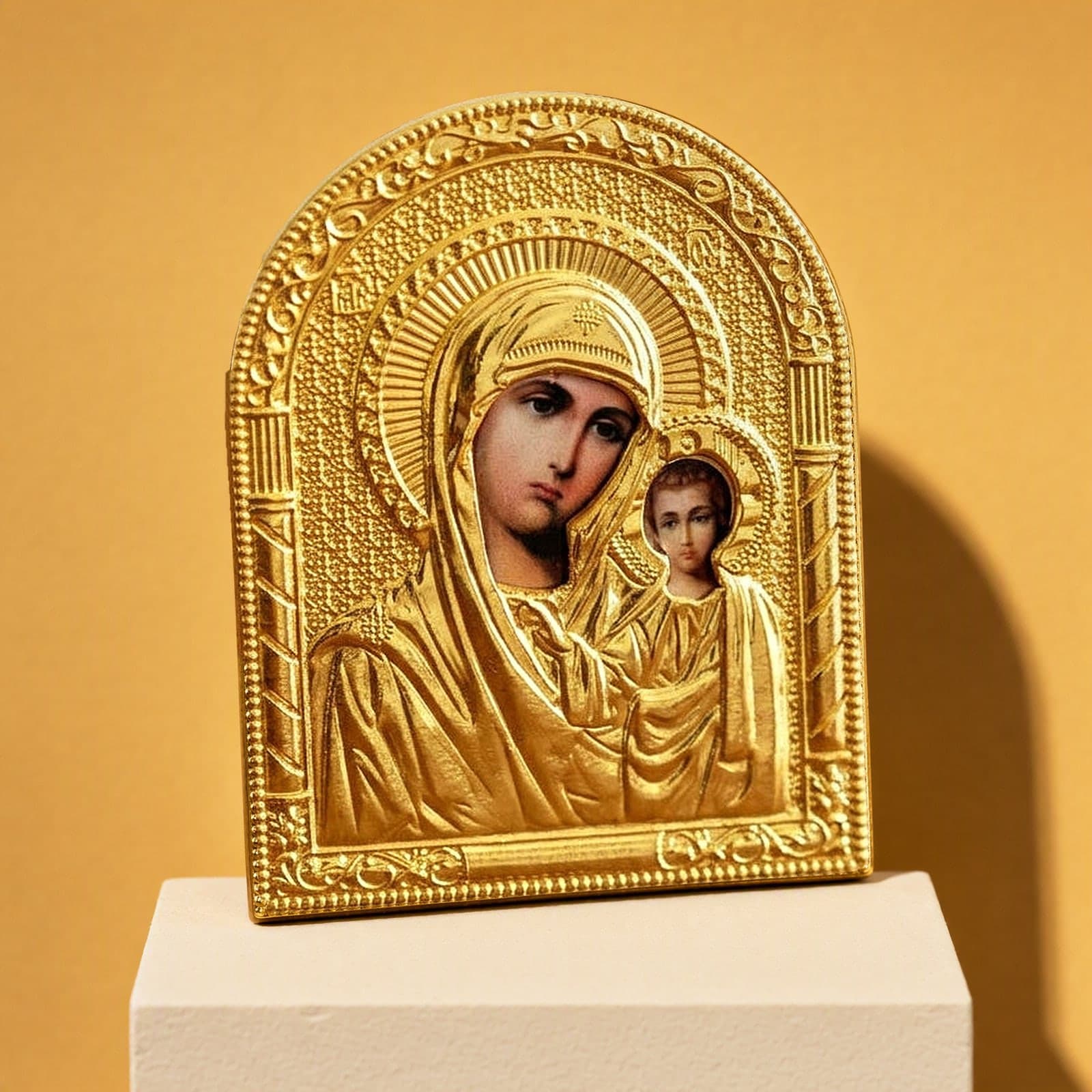 Orthodox Virgin Mary Holding Jesus - Gold Color - Icon: an arched gold-foil icon of the Mother of God of Kazan with the Christ Child, on a white plinth against an ochre backdrop.