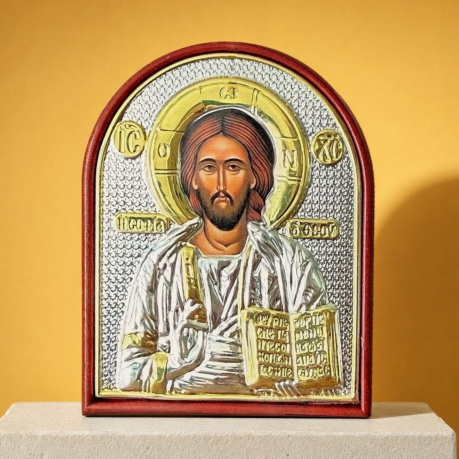 Orthodox Jesus Christ Pantocrator - Silver&Gold Color - Icon: an arched silver-and-gold foil icon of Christ Pantocrator mounted on a wooden board with a red rim, on a white plinth against an ochre backdrop.