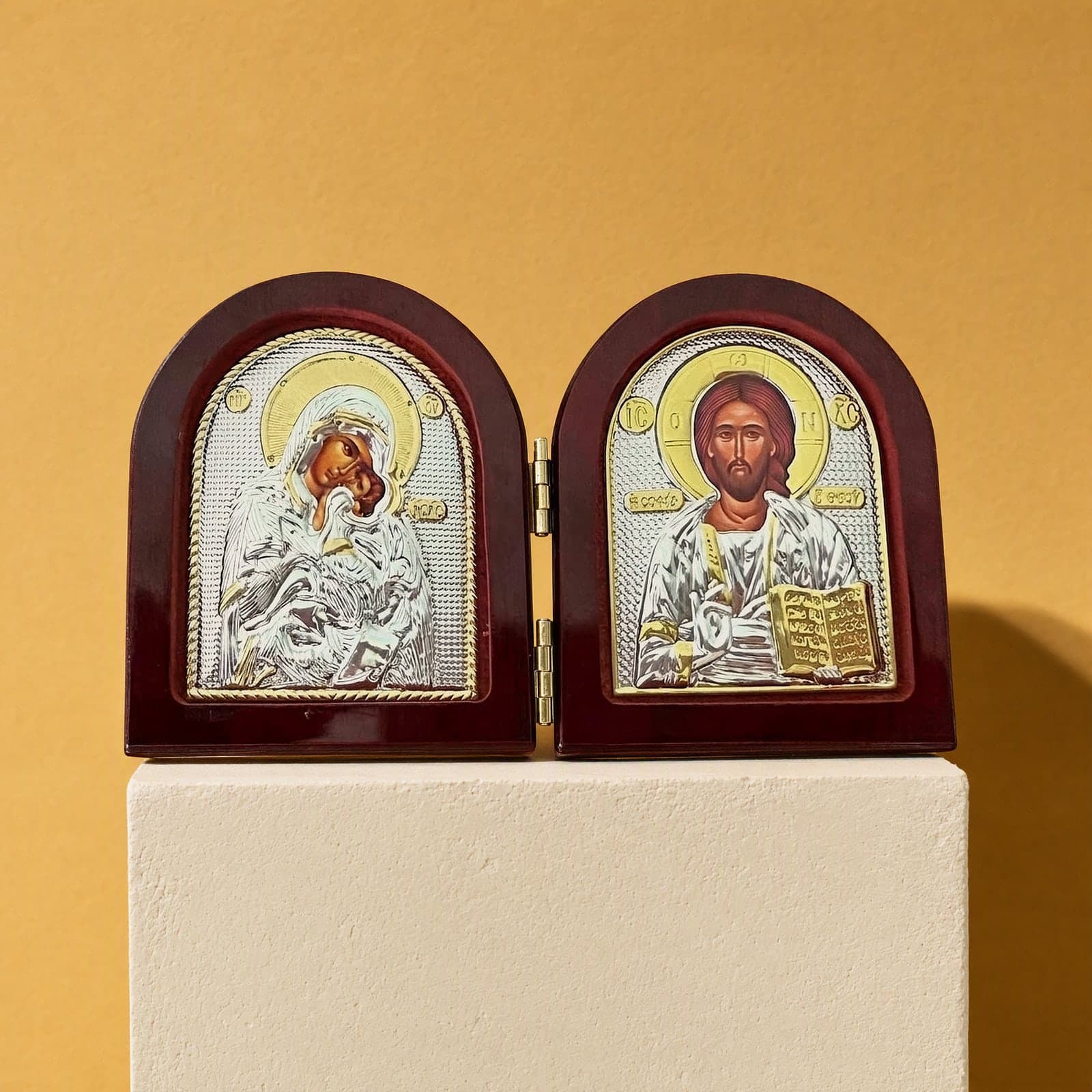 Orthodox Christ & Virgin Mary - Silver&Gold Color - Folding Wooden Icon: a small folding arched icon diptych with silver-and-gold foil icons of the Mother of God and Christ in dark wooden frames, on a white plinth against an ochre backdrop.