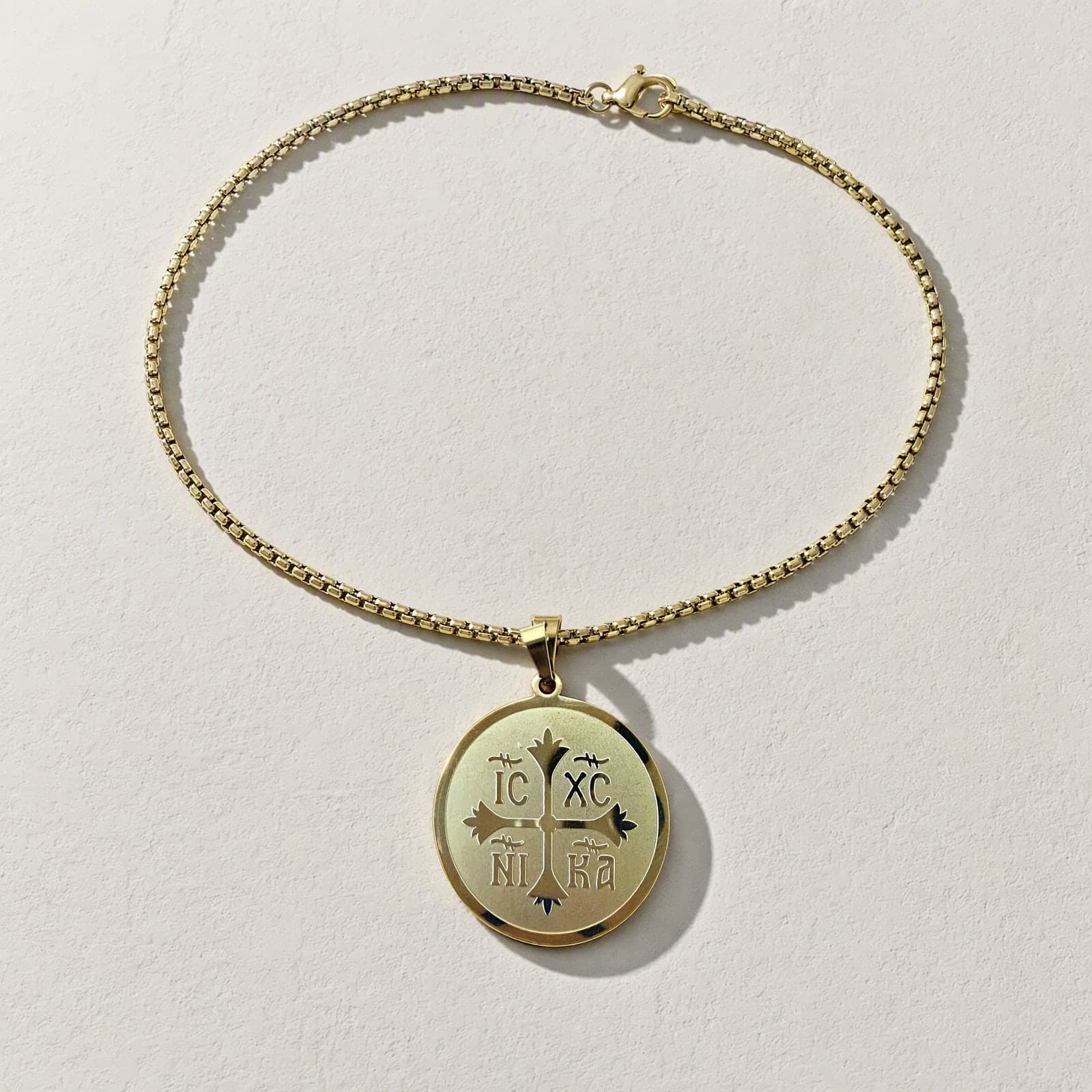 Orthodox IC XC NIKA - Gold Color - Stainless Steel Medallion laid flat with its gold-tone box chain on a white plaster surface.