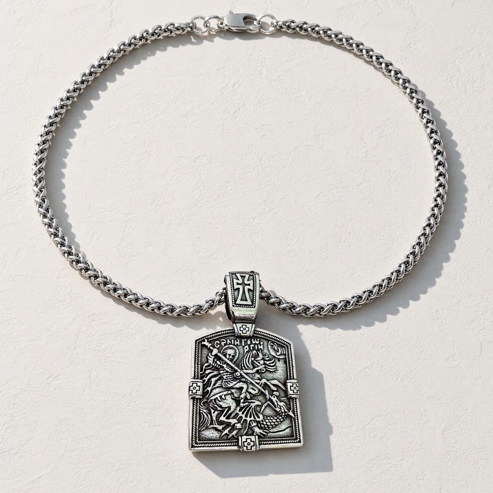 Orthodox St. George - Silver Color - 60cm Chain - Stainless Steel Medallion laid flat with its silver-tone wheat chain on a white plaster surface.