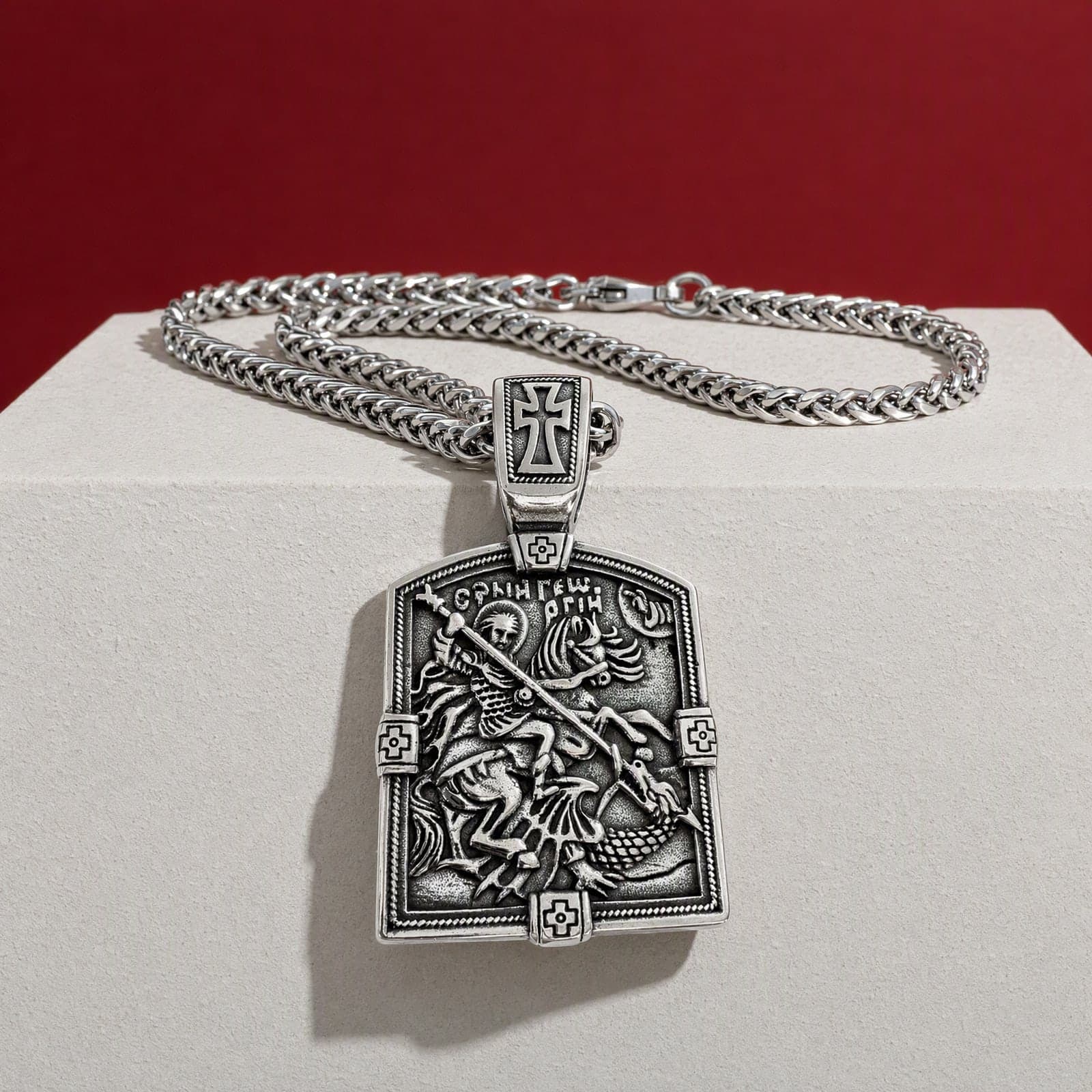 Orthodox St. George, silver-tone stainless steel medallion, hanging over the edge of a bone plaster plinth against an oxblood backdrop, on a silver-tone wheat chain.