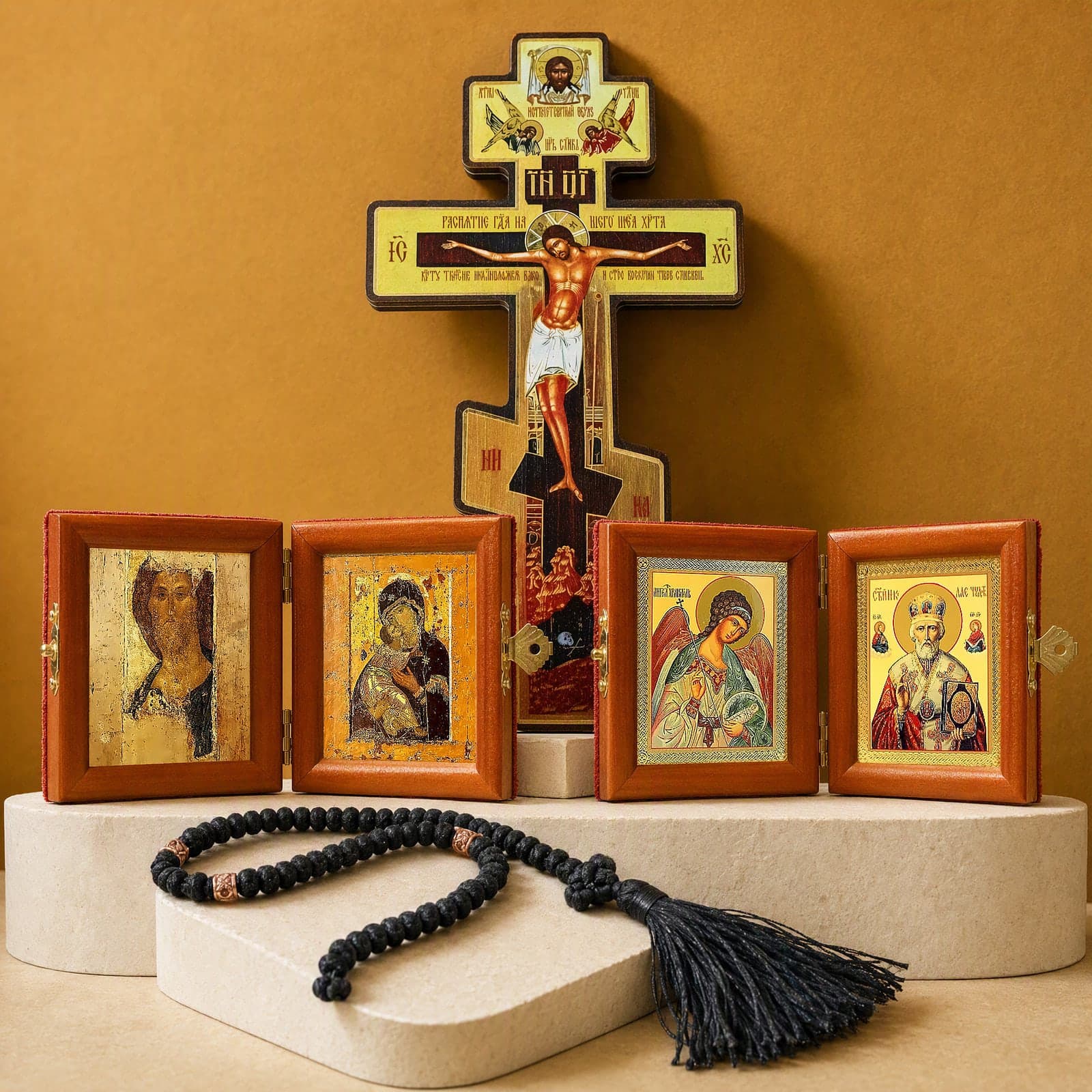 Icon Corner Set: the folding diptych of Christ in the Rublev manner (the Zvenigorod Saviour) on an aged gold ground and the Vladimir Mother of God holding the Christ Child cheek to cheek; the folding diptych of the Guardian Angel holding a cross and an orb and St Nicholas the Wonderworker in a jewelled mitre holding the Gospel (one of the patron saints you can choose); an 8 inch wooden Orthodox wall crucifix with a painted image of the Crucifixion; a black wool Orthodox prayer rope of 50 knots with copper-tone wooden beads, a knotted cross and a black tassel; on bone plaster plinths against an ochre backdrop (icon-corner guide not shown).