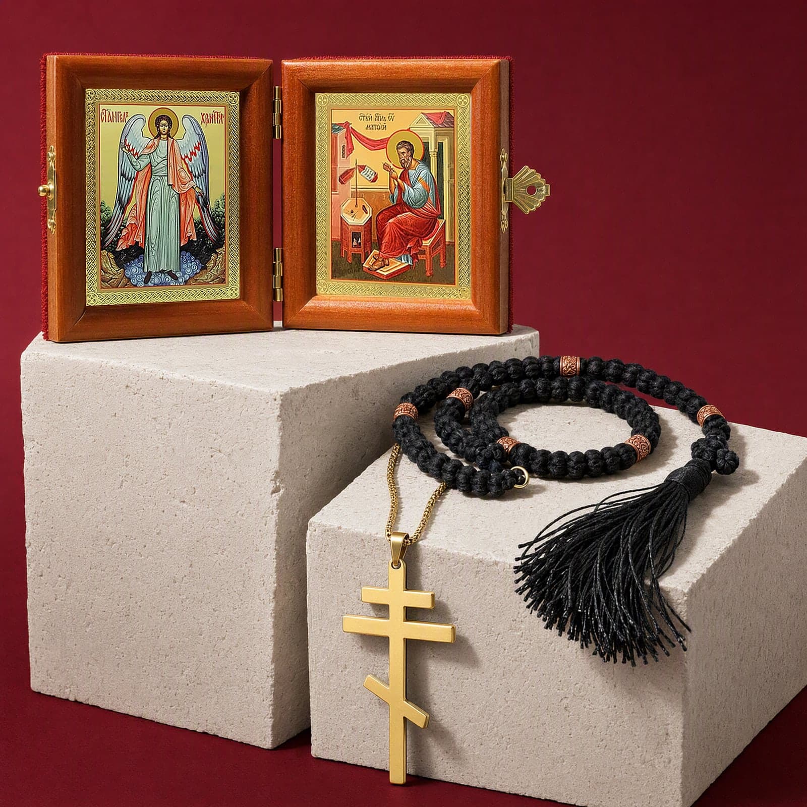Godparent Set: the folding diptych of the Guardian Angel standing full length and St Matthew the Apostle writing his Gospel (one of the patron saints you can choose); a plain polished gold-tone stainless steel three-bar Orthodox cross with the slanted foot bar on a gold-tone box chain; a black wool Orthodox prayer rope of 50 knots with copper-tone wooden beads, a knotted cross and a black tassel; on bone plaster plinths against an oxblood backdrop (holy-water vial and date card not shown).