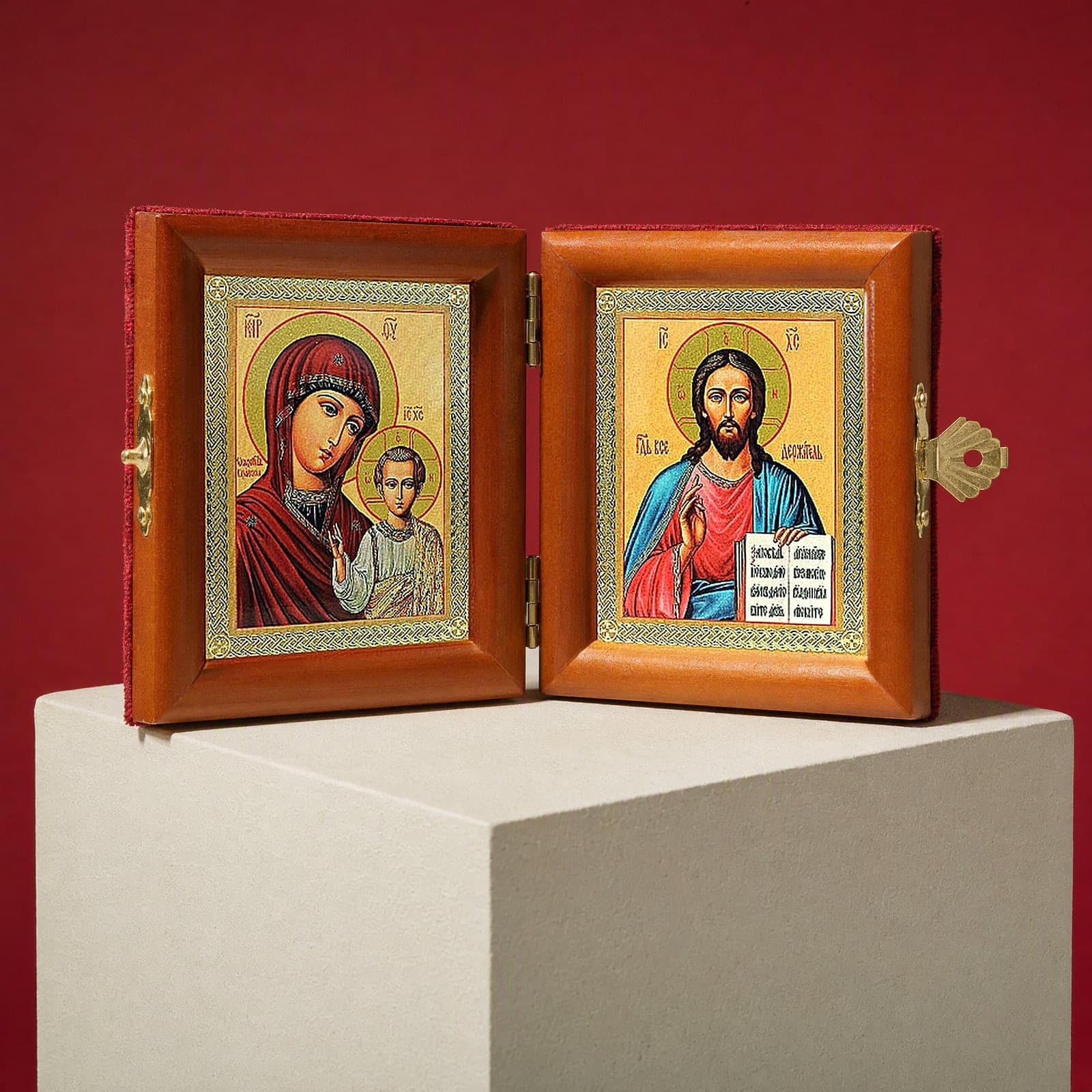 Christ Pantocrator & Our Lady of Kazan travel diptych seen from a 45-degree angle, half open on a white plinth against an oxblood backdrop, showing the frame depth and hinges.