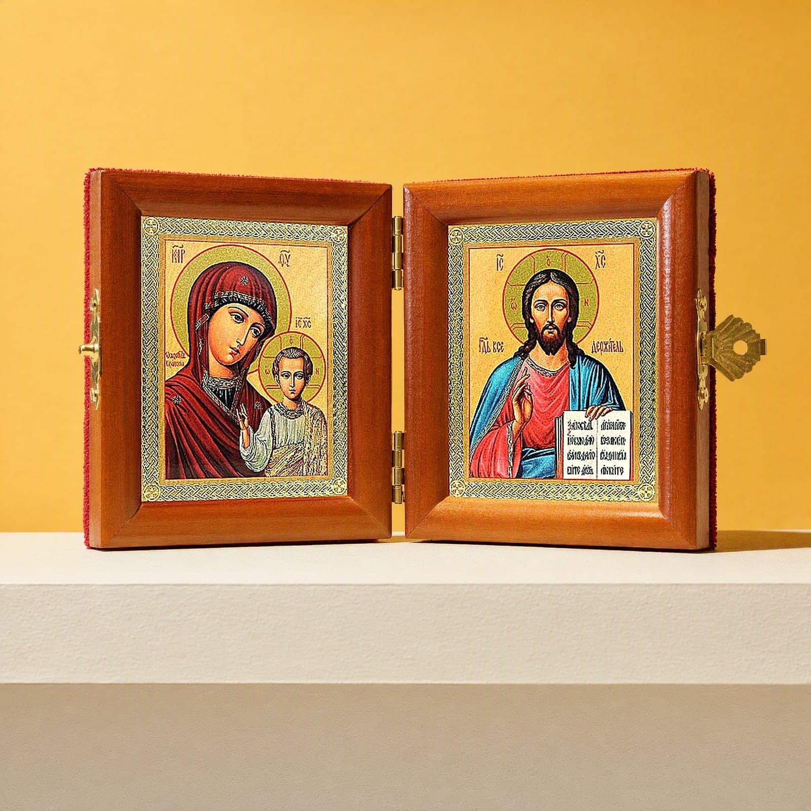 Christ Pantocrator & Our Lady of Kazan travel diptych, open: folding diptych of Our Lady of Kazan with the Christ Child and Christ Pantocrator holding an open Gospel in honey pine frames with brass hinges and clasp, on a white plinth against an ochre backdrop.