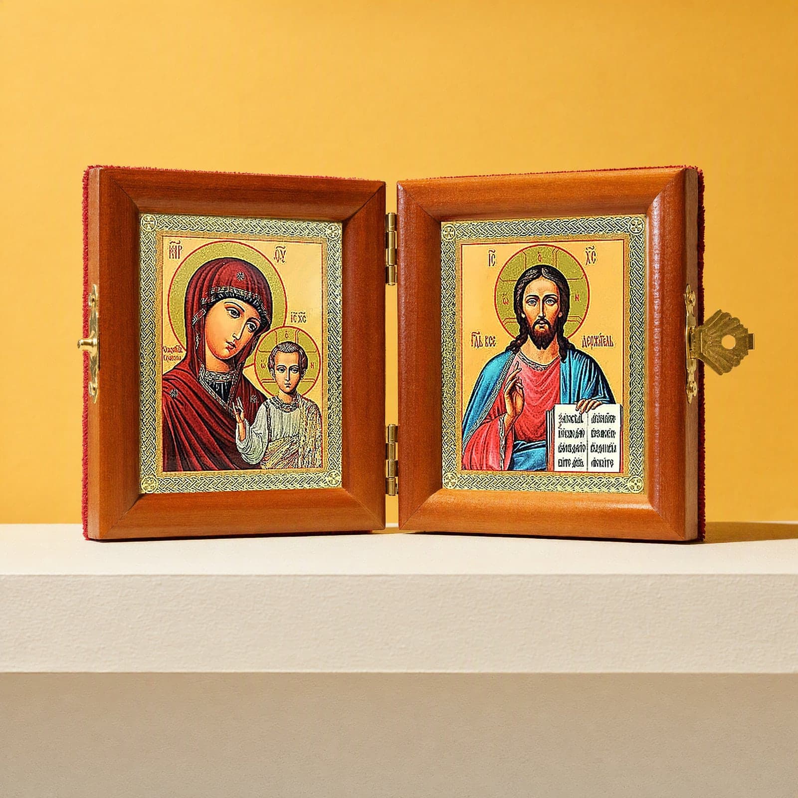 Christ Pantocrator & Our Lady of Kazan travel diptych, open: folding diptych of Our Lady of Kazan with the Christ Child and Christ Pantocrator holding an open Gospel in honey pine frames with brass hinges and clasp, on a white plinth against an ochre backdrop.