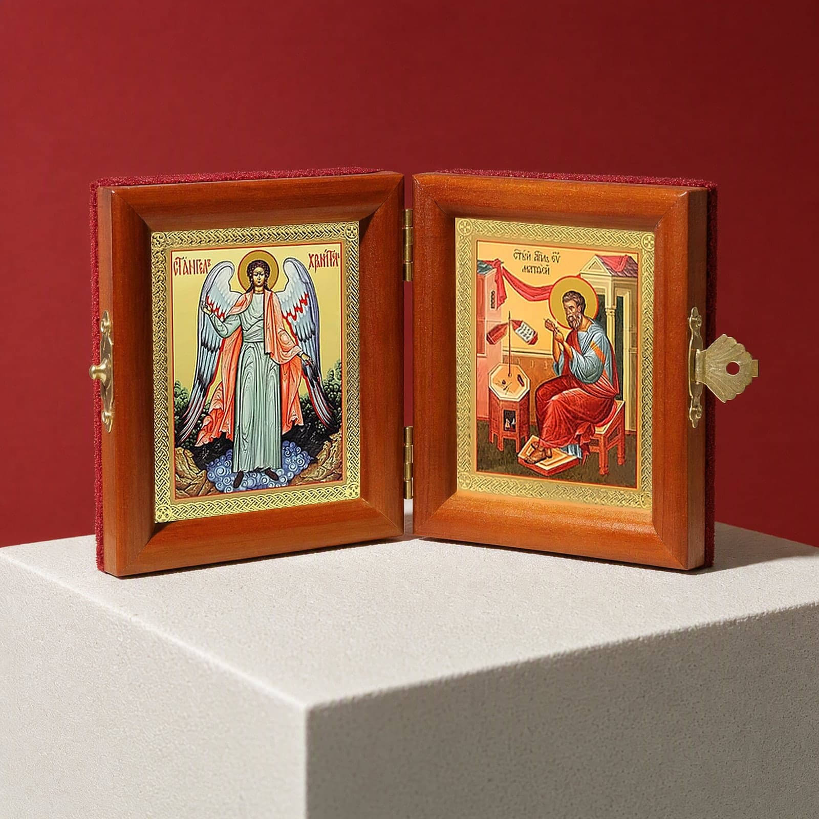 St Matthew the Apostle & Guardian Angel name diptych seen from a 45-degree angle, half open on a white plinth against an oxblood backdrop, showing the frame depth and hinges.