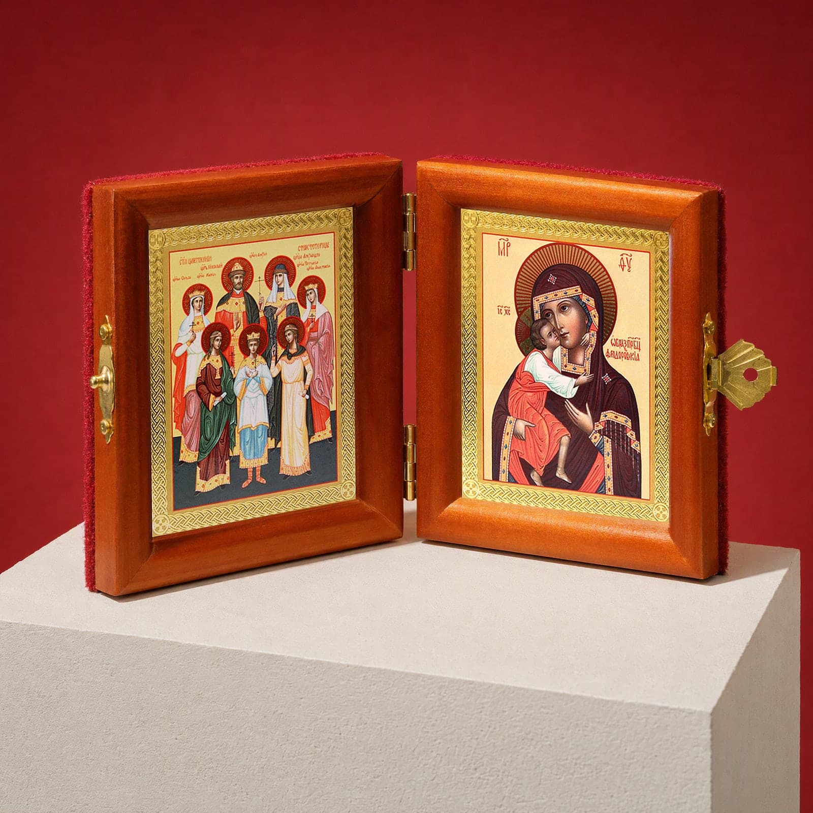 Royal Passion-Bearers (Romanov family) & Feodorovskaya Mother of God travel diptych seen from a 45-degree angle, half open on a white plinth against an oxblood backdrop, showing the frame depth and hinges.