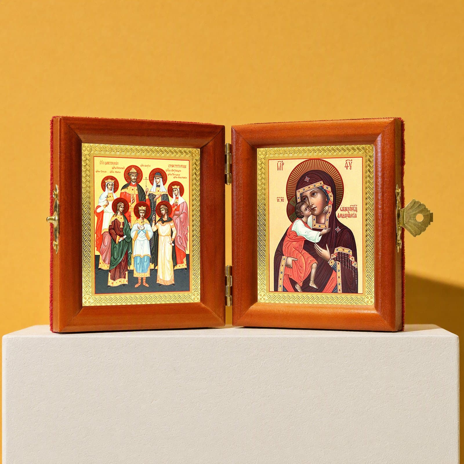 Royal Passion-Bearers (Romanov family) & Feodorovskaya Mother of God travel diptych, open: folding diptych of the Royal Passion-Bearers, the family of Tsar Nicholas II and the Feodorovskaya Mother of God in honey pine frames with brass hinges and clasp, on a white plinth against an ochre backdrop.
