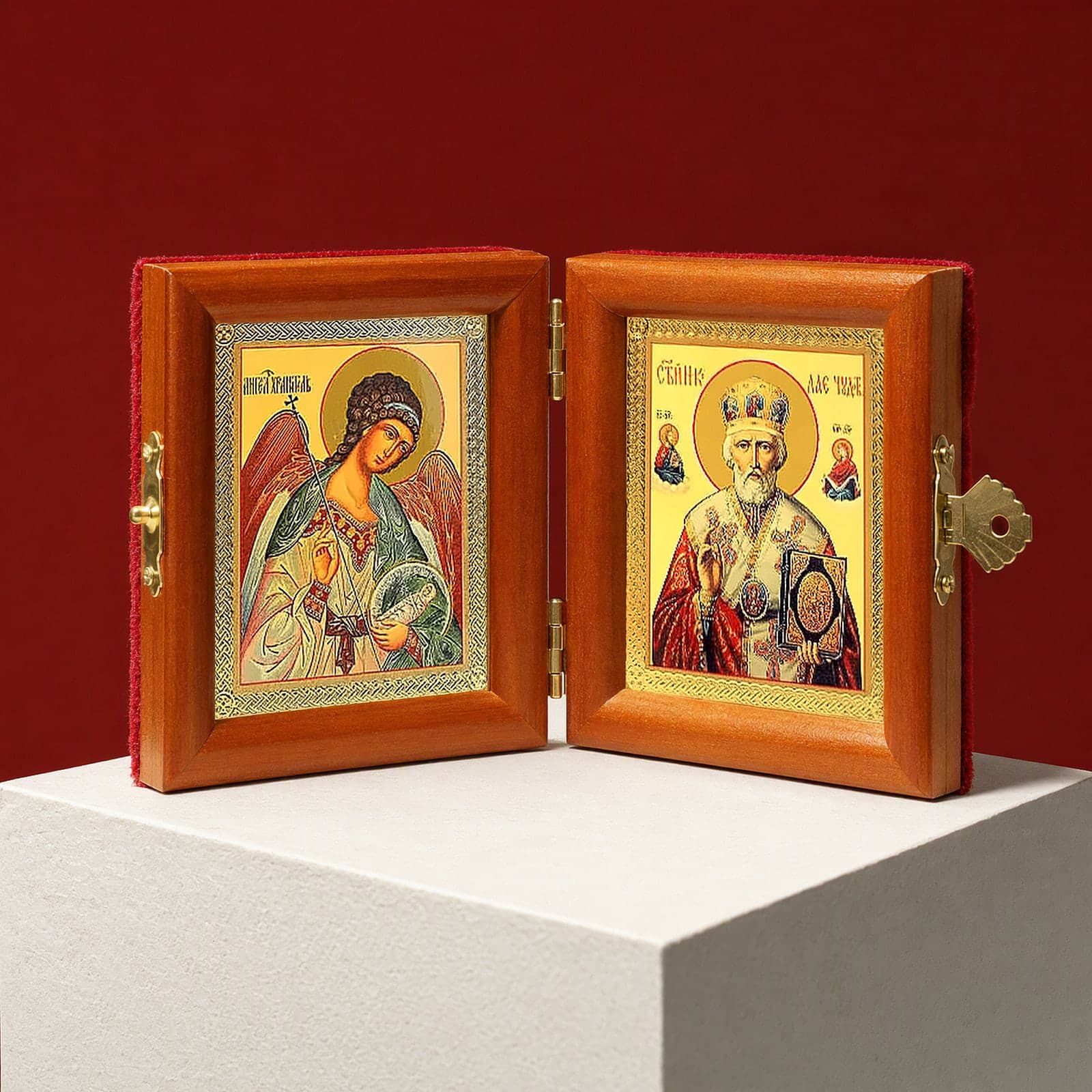 St Nicholas the Wonderworker & Guardian Angel name diptych seen from a 45-degree angle, half open on a white plinth against an oxblood backdrop, showing the frame depth and hinges.